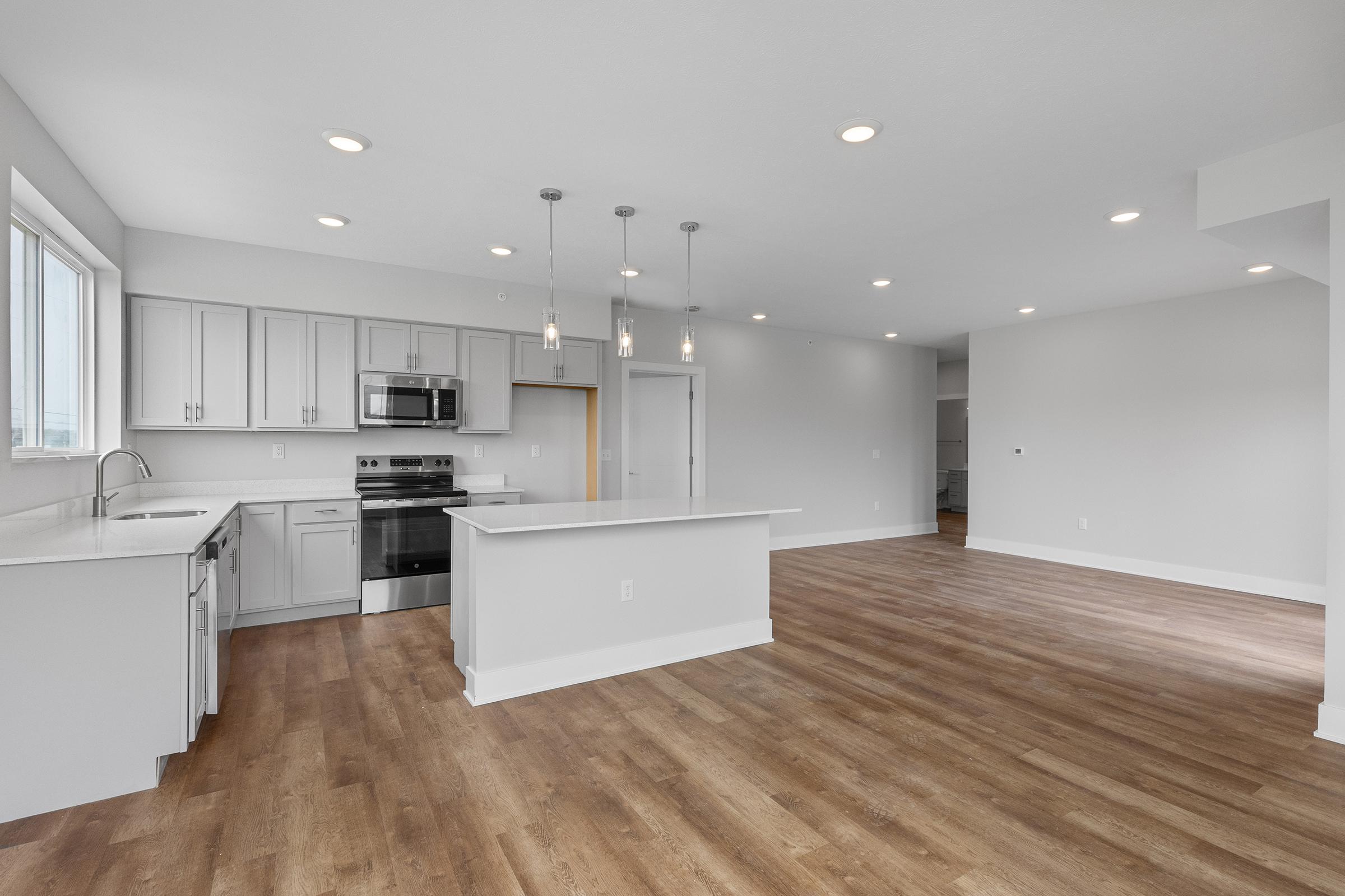 Modern open-concept kitchen and living area featuring light gray cabinetry, stainless steel appliances, and a large island with seating. The space is well-lit with pendant lighting and large windows, showcasing a clean, minimalist design with warm wood flooring.