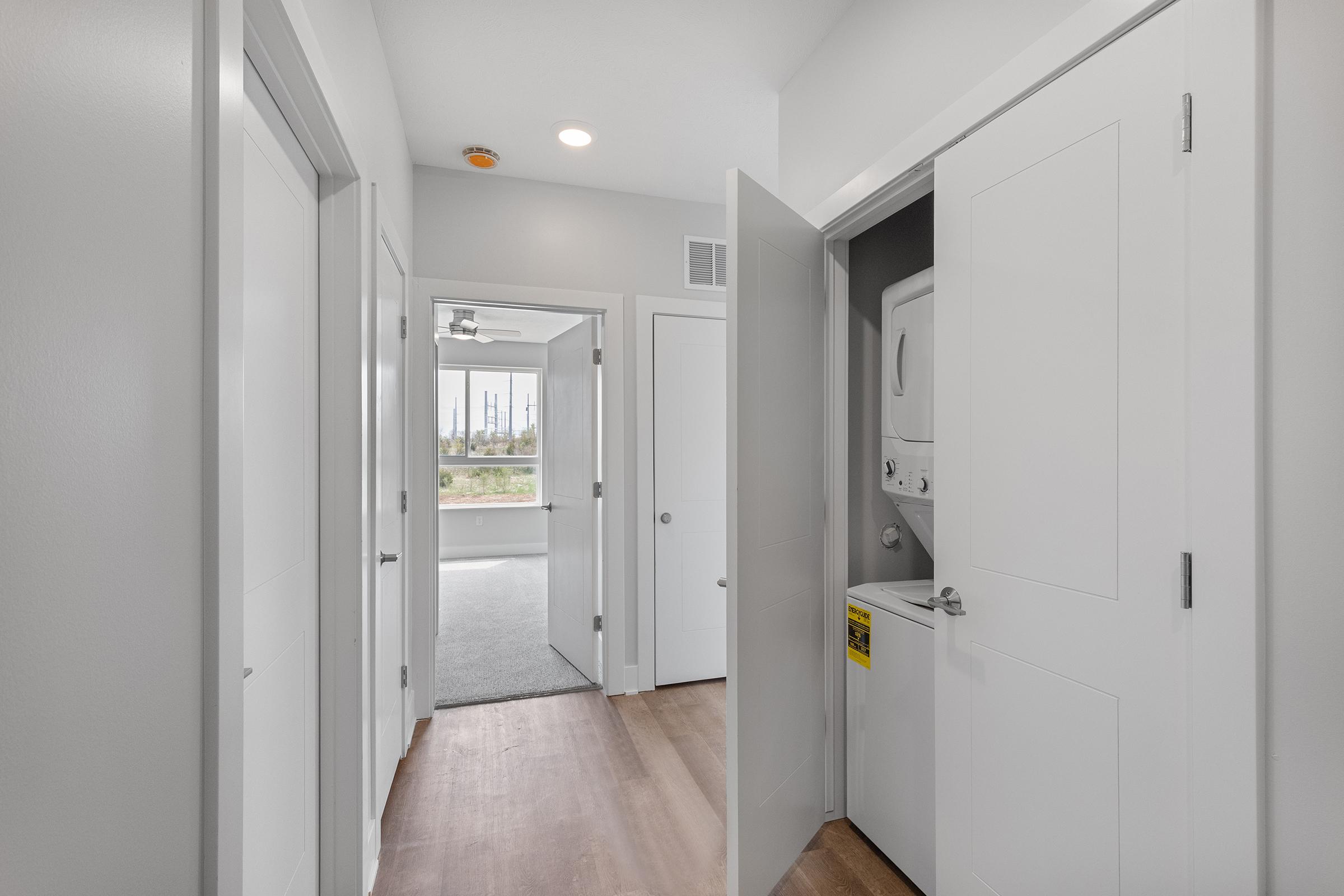 A bright, modern hallway with light gray walls and a hardwood floor. Two doors are open: one leads to a laundry area with a stacked washer and dryer, and the other opens to a spacious room with large windows. Natural light fills the space, enhancing the airy atmosphere.