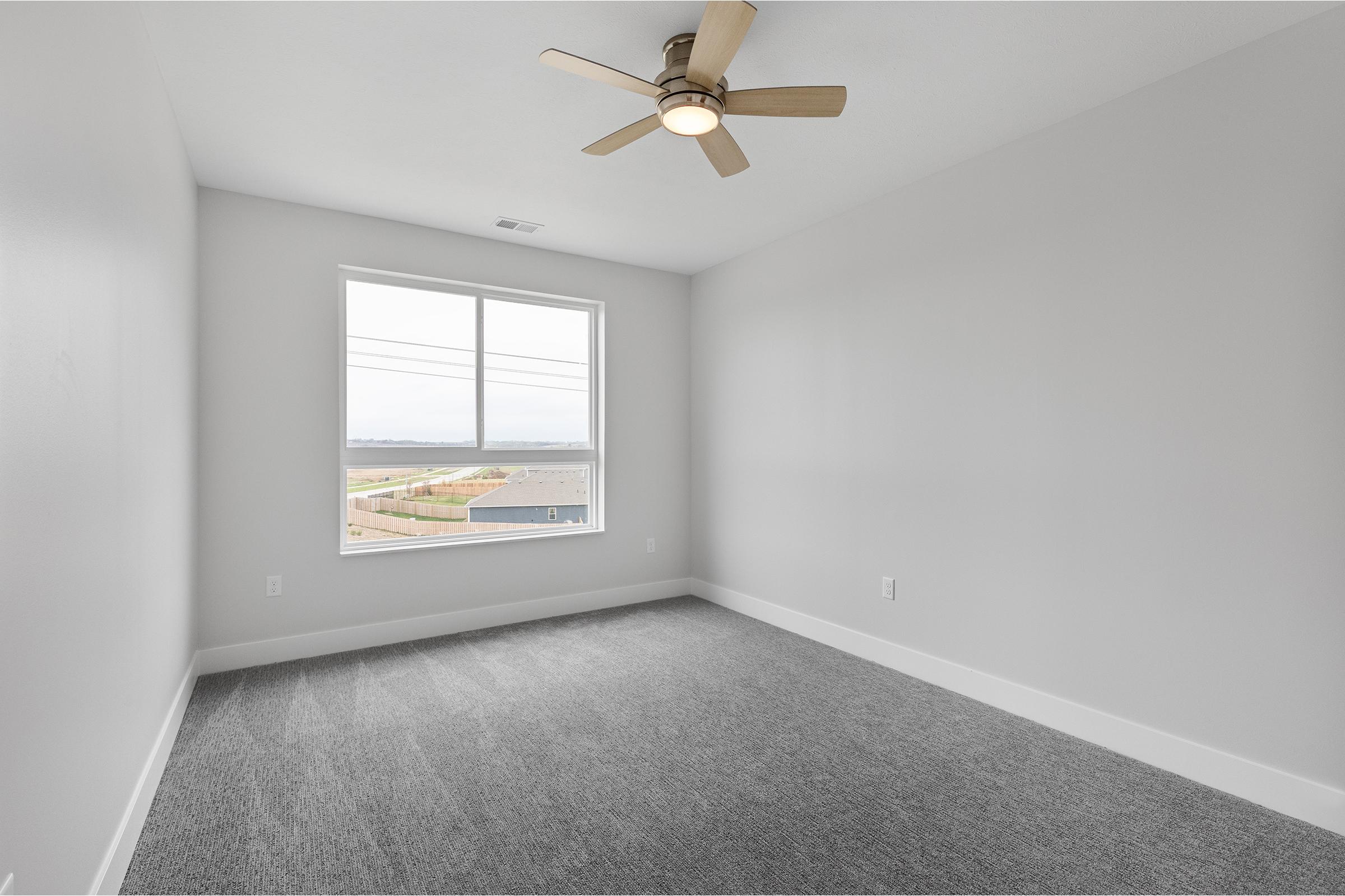 A spacious, empty bedroom featuring light gray walls and a ceiling fan. There is a large window providing natural light and a view of the outside landscape. The floor is covered with gray carpet, creating a clean and modern atmosphere.