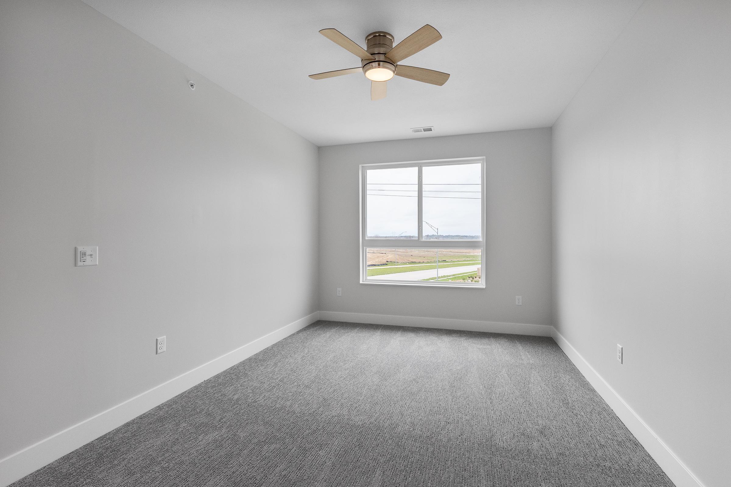 Empty room with light gray walls and carpet, featuring a ceiling fan and a large window allowing natural light. The view outside includes a grassy area and distant wind turbines.