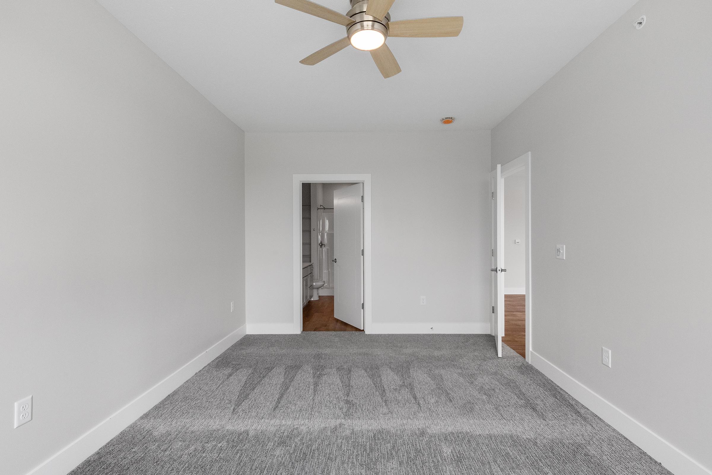 A spacious, empty room featuring light gray walls and plush gray carpeting. A ceiling fan is mounted above, and there are two doorways leading to adjacent areas, one possibly to a bathroom. Natural light brightens the space, creating a modern and inviting atmosphere.