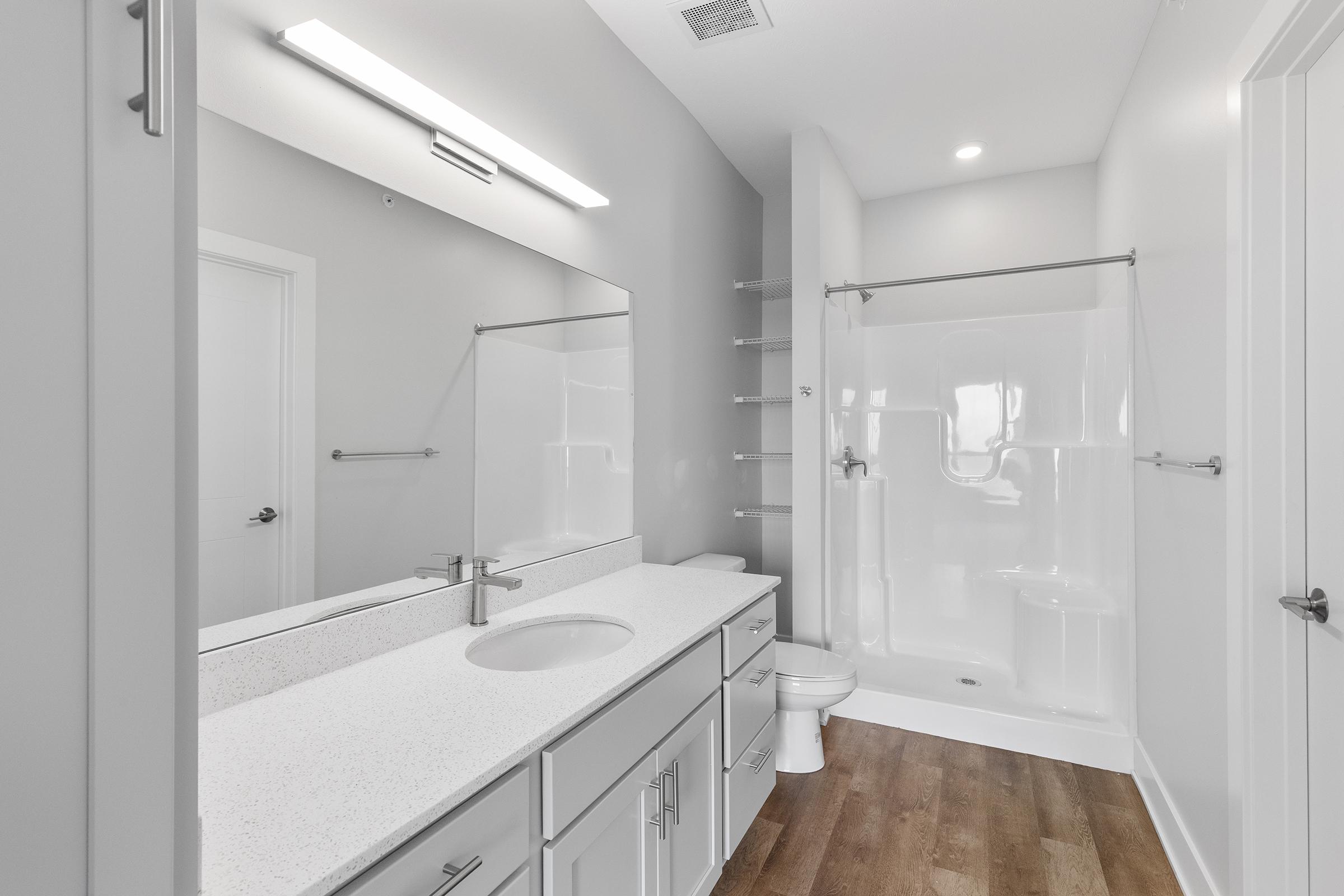 A modern bathroom featuring a spacious vanity with a sink, large mirror, and contemporary fixtures. The shower area has a glass door and a white tub, with light-colored walls and wooden flooring, creating a clean and minimalistic aesthetic. Good natural and artificial lighting enhance the space.