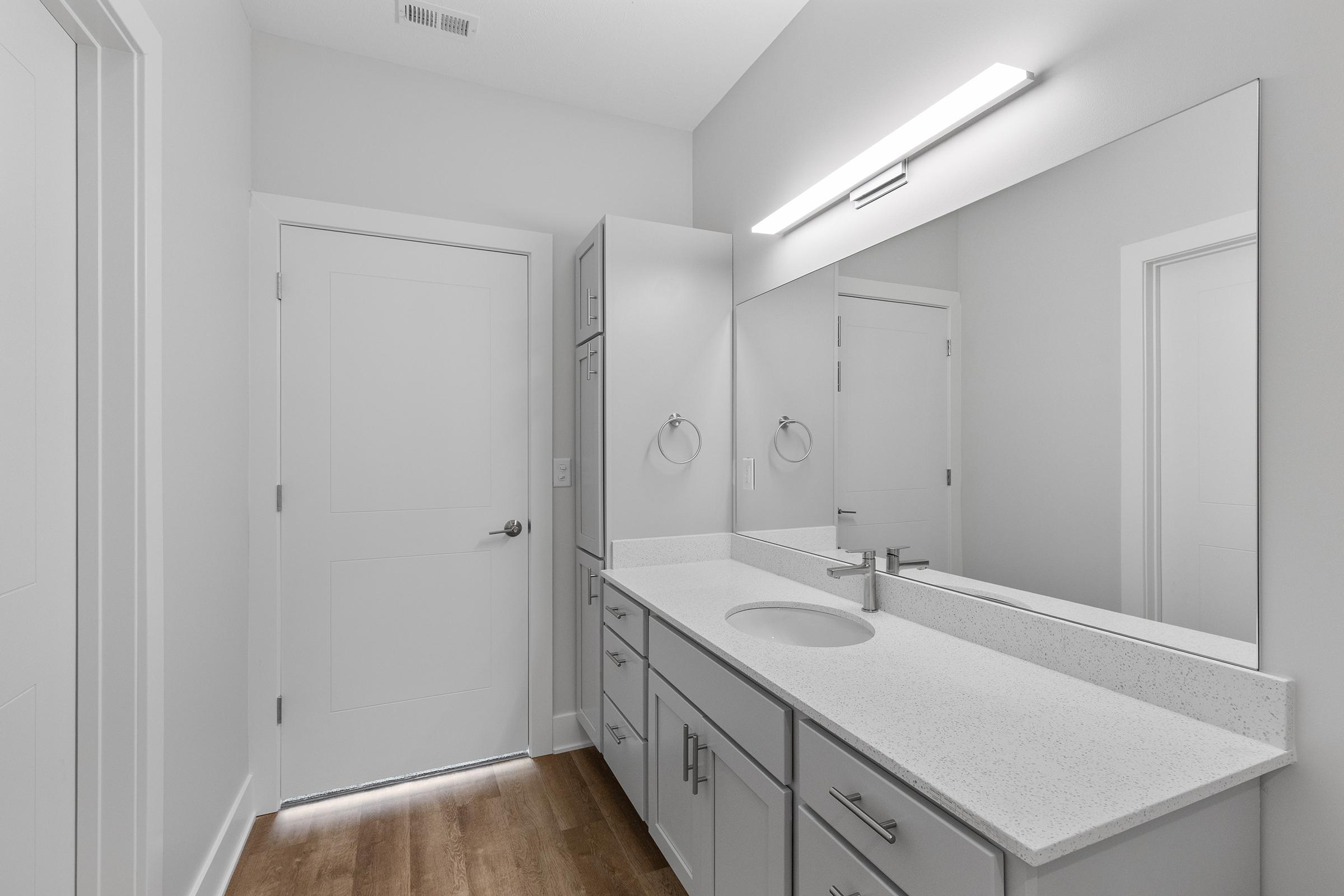 Modern bathroom featuring a sleek vanity with a white countertop and single sink, large mirror above, and gray cabinets. The floor is hardwood, and there are white walls and a door in the background. A bright light fixture is mounted above the mirror, providing ample illumination.