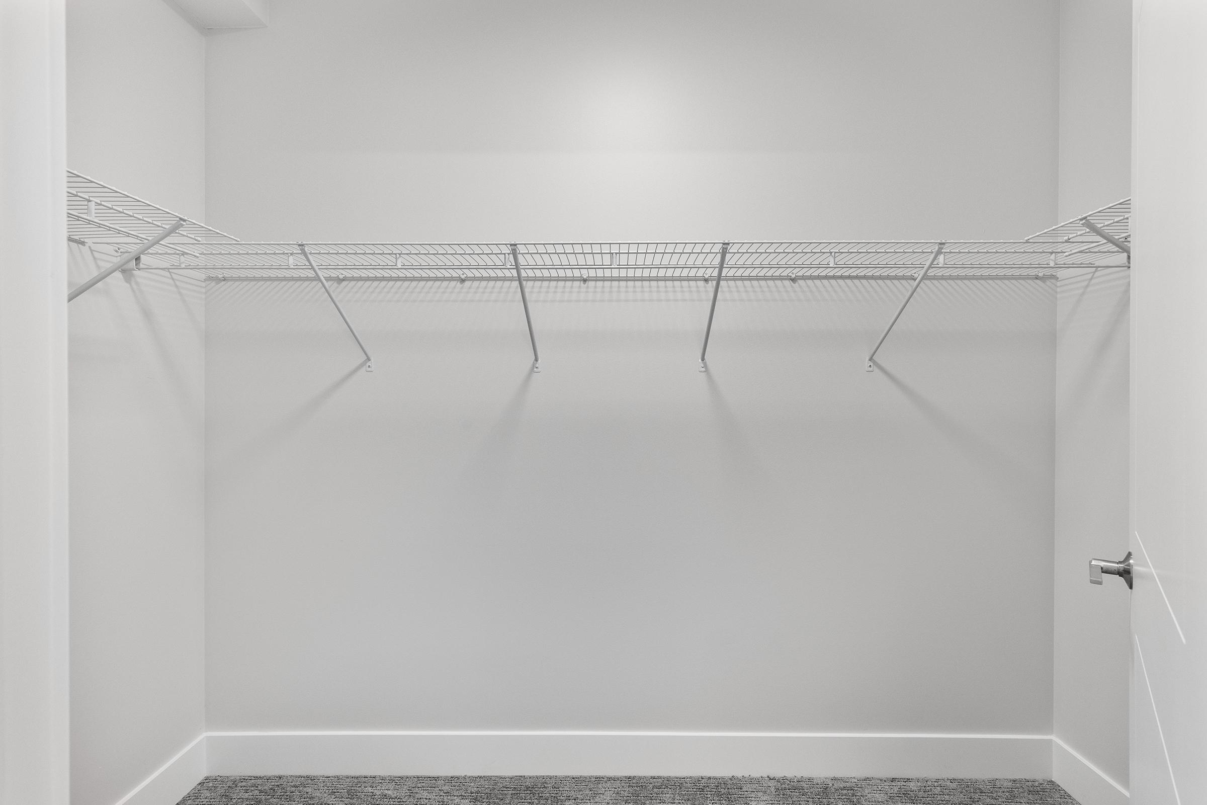 Empty closet with white walls and a wire shelving unit mounted on one side. The floor is covered with gray carpet, and there is a white door partially visible on the right. The space is well-lit, emphasizing its emptiness.