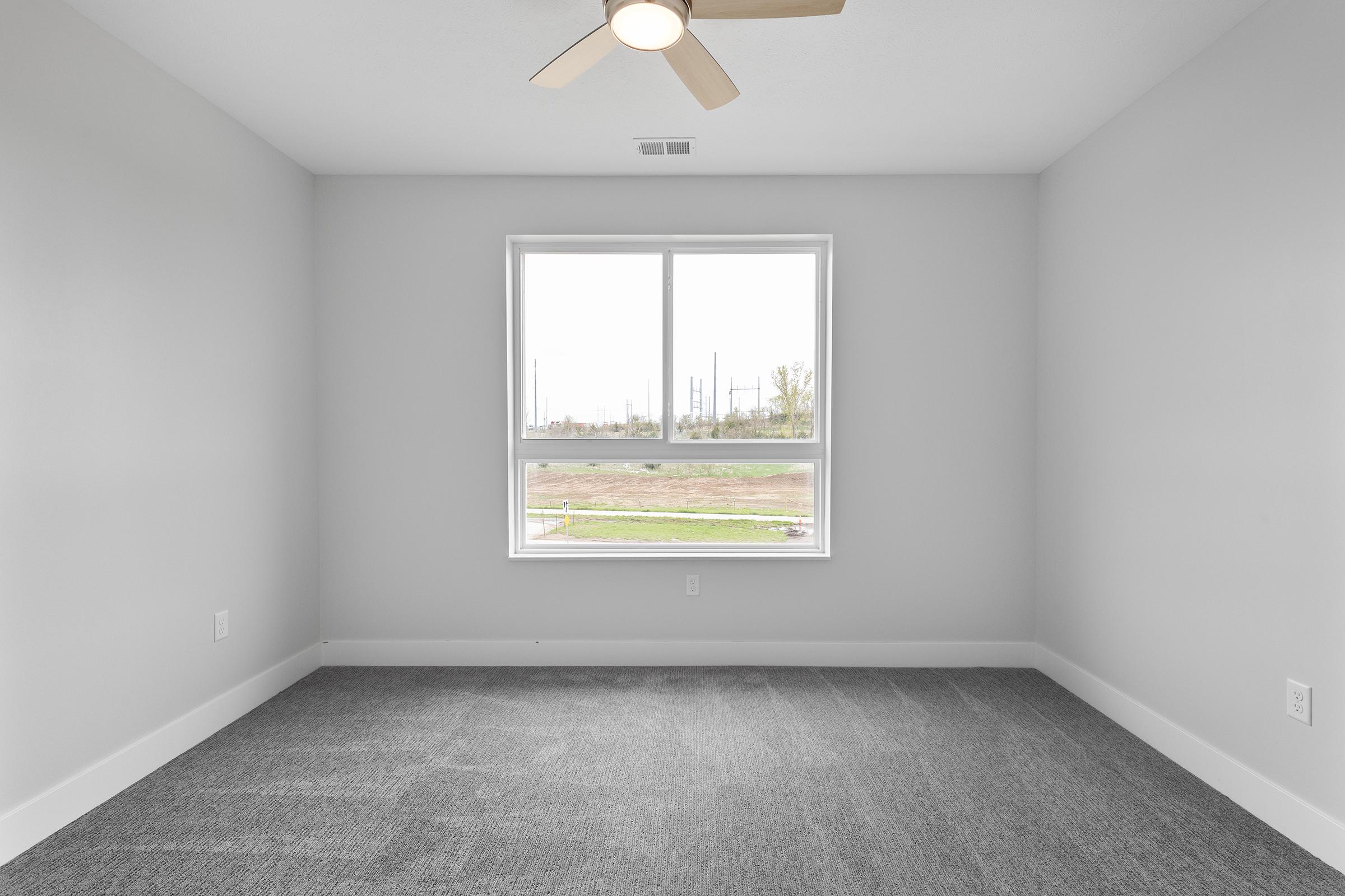 A blank, minimalist room with gray carpeting and light gray walls. A large window lets in natural light, showing an outdoor view with trees and utility poles. The ceiling features a light-colored ceiling fan. The space appears fresh and ready for furnishing.