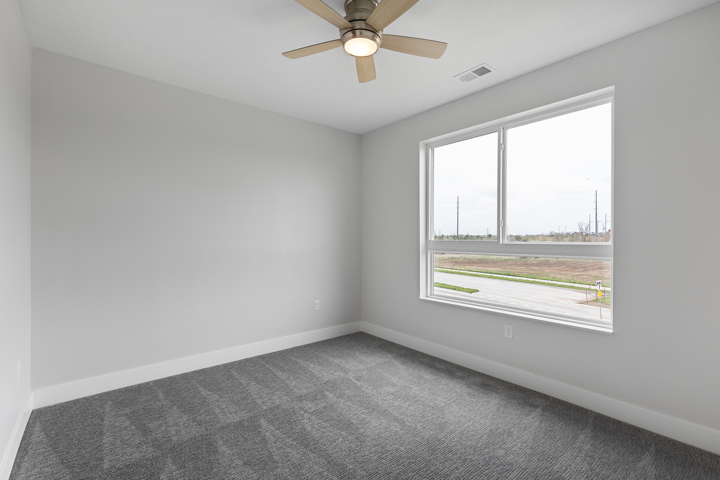 Empty room with light gray walls and a large window, allowing natural light to enter. A ceiling fan is visible, and the floor is covered with dark gray carpet. The view outside shows an open area with fields and distant structures.