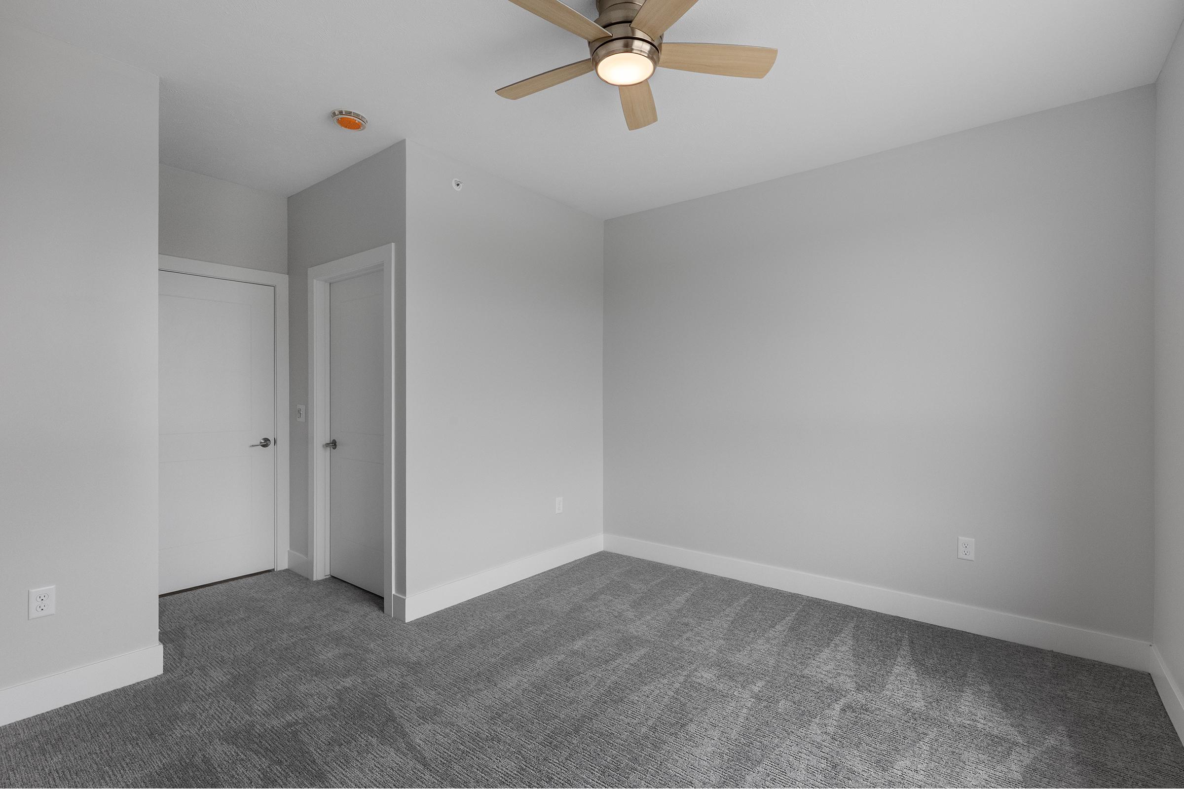 A light gray room with a ceiling fan and new carpet. There is a closed white door on the left and an open white door on the right, leading to another area. The walls are bare and the overall atmosphere is clean and modern, suggesting it could be a bedroom or office space.