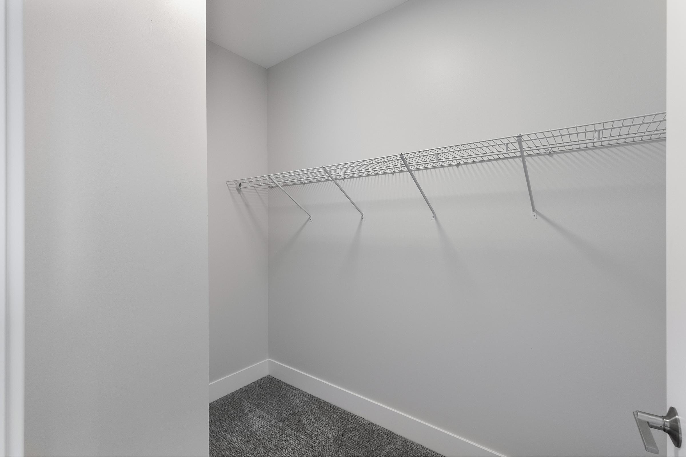 A simple, empty closet with light gray walls. It features a wire shelving unit along one side and a clean, carpeted floor. The door to the closet is partially open, allowing a view of the minimalistic space.