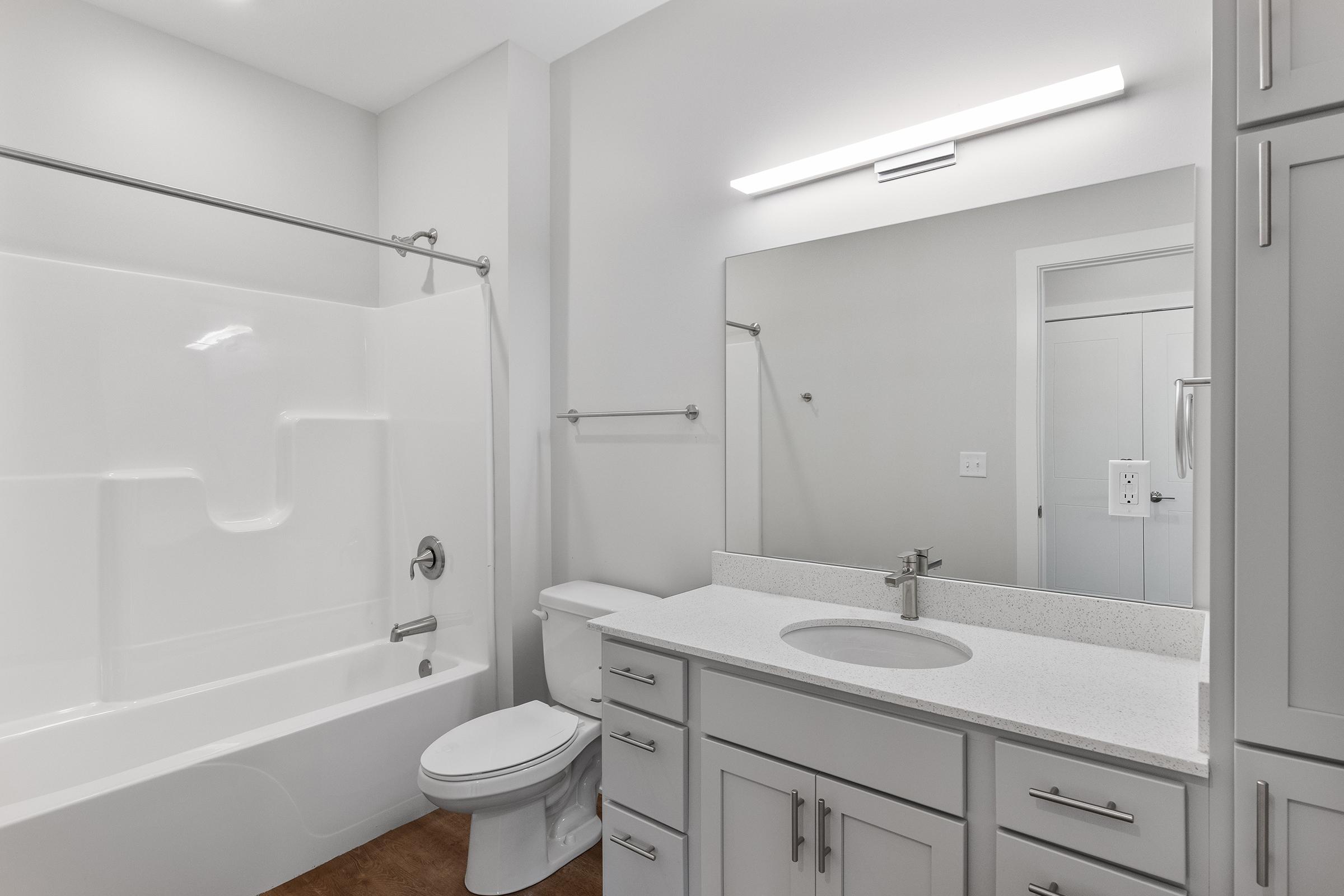 A modern bathroom featuring a white bathtub with a shower, a sleek white vanity with a sink, and a large mirror above it. The walls are painted in neutral tones, and the floor has a warm wood finish. A towel rack is mounted next to the bathtub, and the overall design is clean and minimalist.