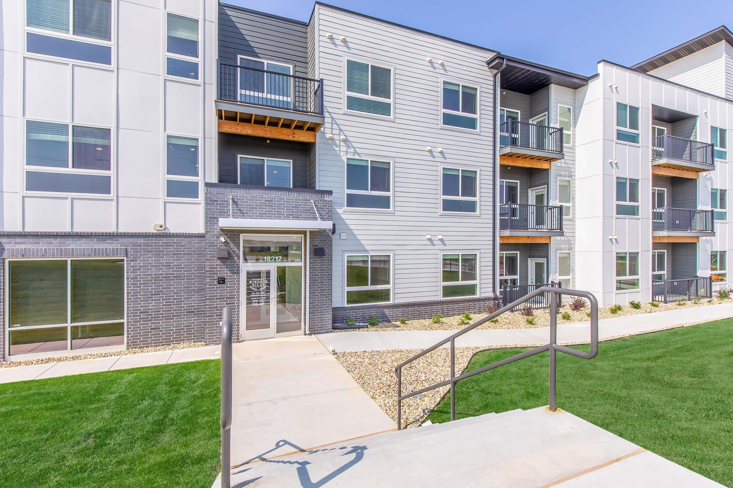 Modern apartment building exterior featuring a combination of gray and white siding, large windows, and balconies. The entrance is accessible via a ramp, with a landscaped lawn and walkways. Bright sunny day enhances the building's fresh, contemporary appearance.