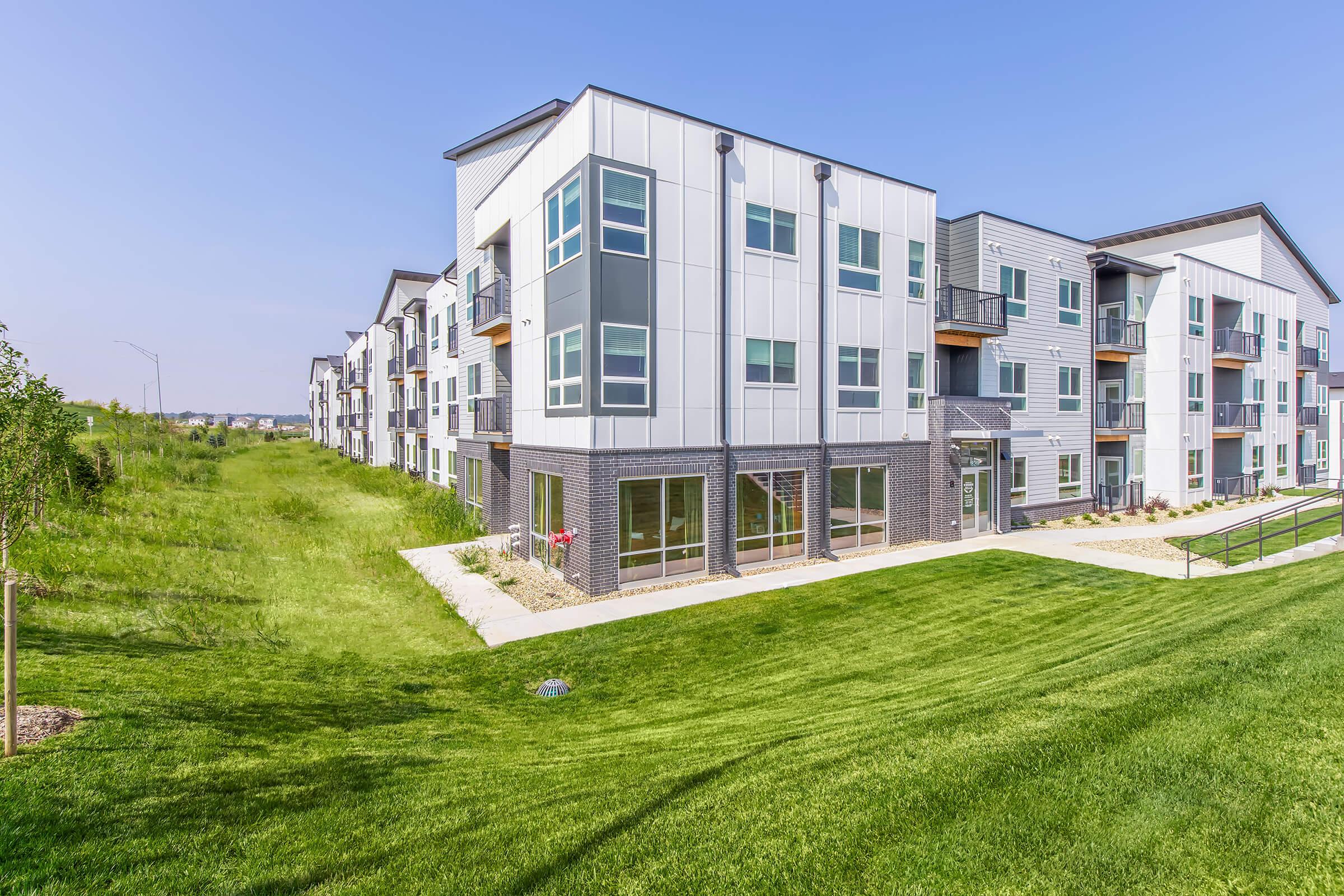 A modern multi-unit residential building with a sleek design, featuring large windows and balconies. The structure is surrounded by neatly maintained green lawns and landscaping, with a clear blue sky in the background, suggesting a bright and inviting atmosphere.