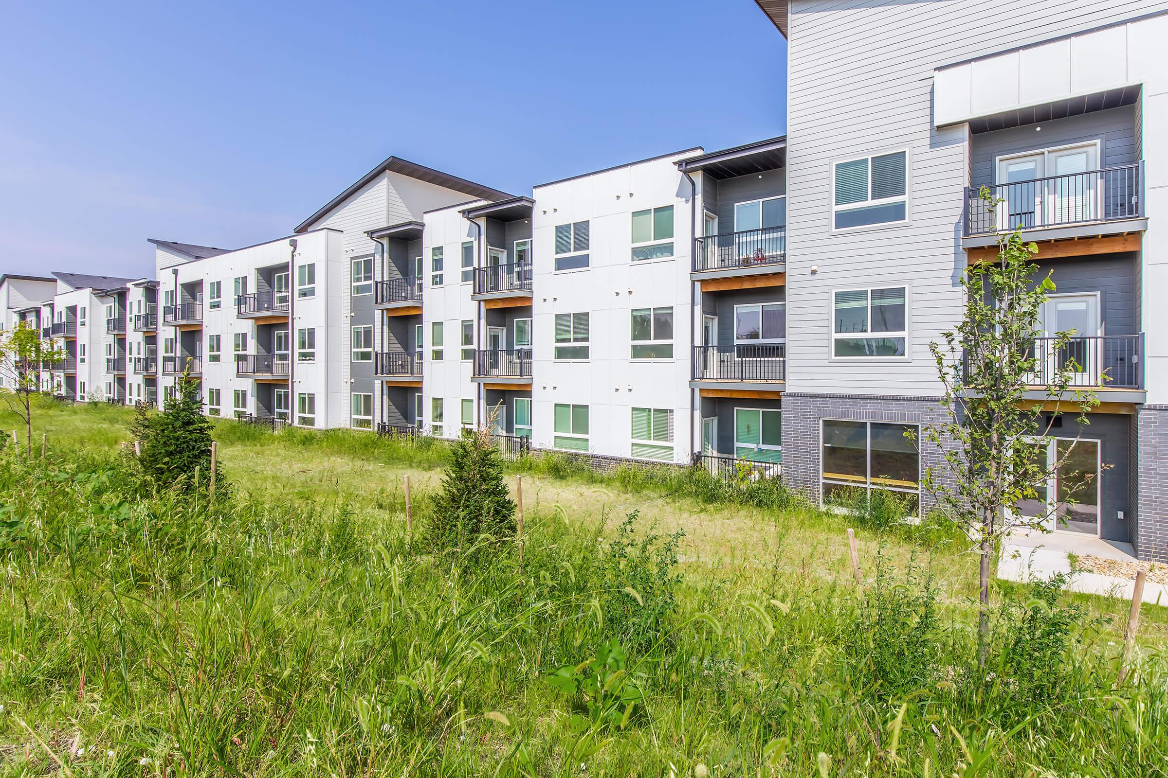 Modern apartment buildings with multiple balconies, surrounded by green grass and sparse landscaping under a clear blue sky. The architecture features a combination of white and gray exteriors, with large windows and sliding glass doors.
