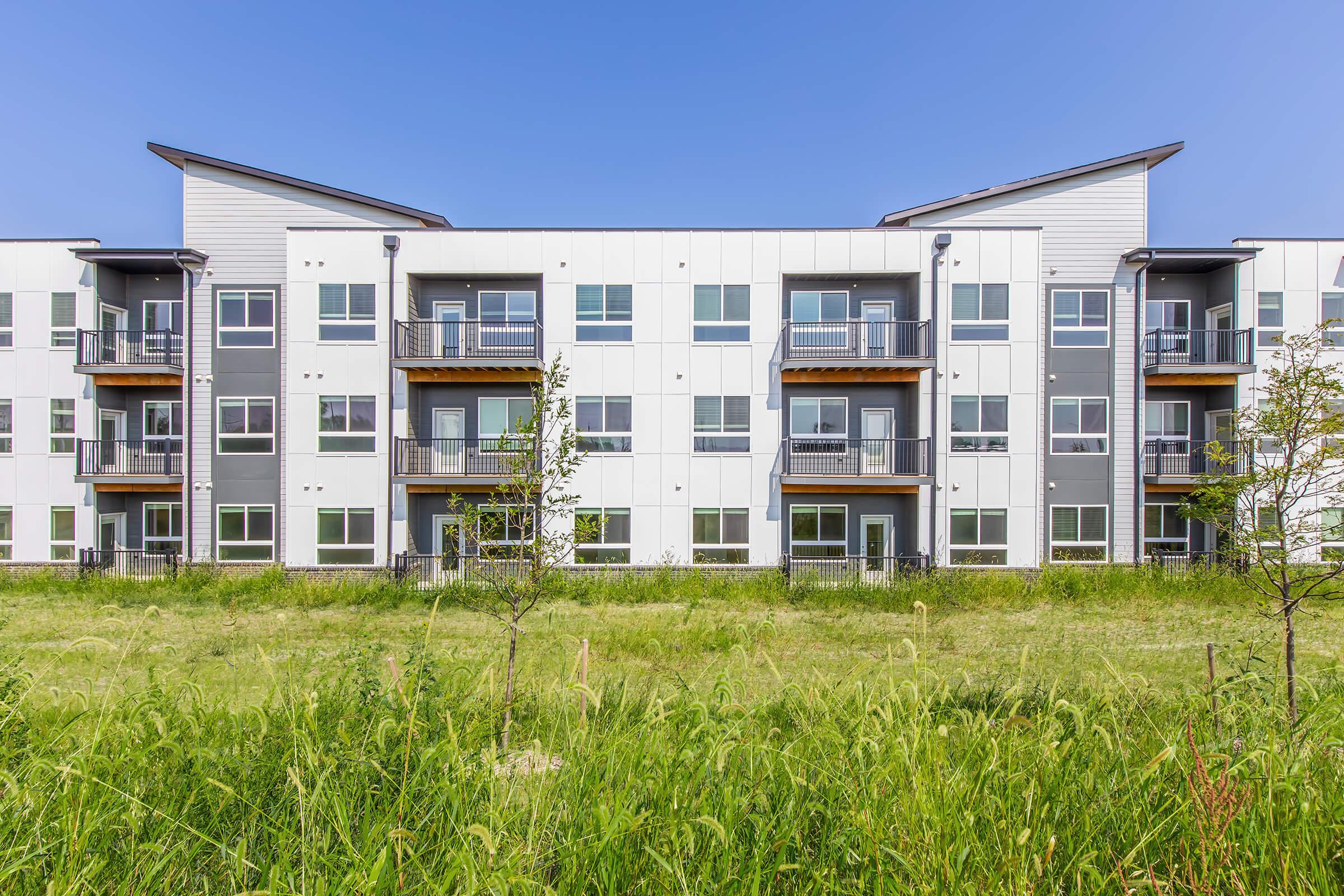 A modern multi-story apartment building with a light and dark gray exterior. The structure features several balconies and large windows, surrounded by a grassy field under a clear blue sky.