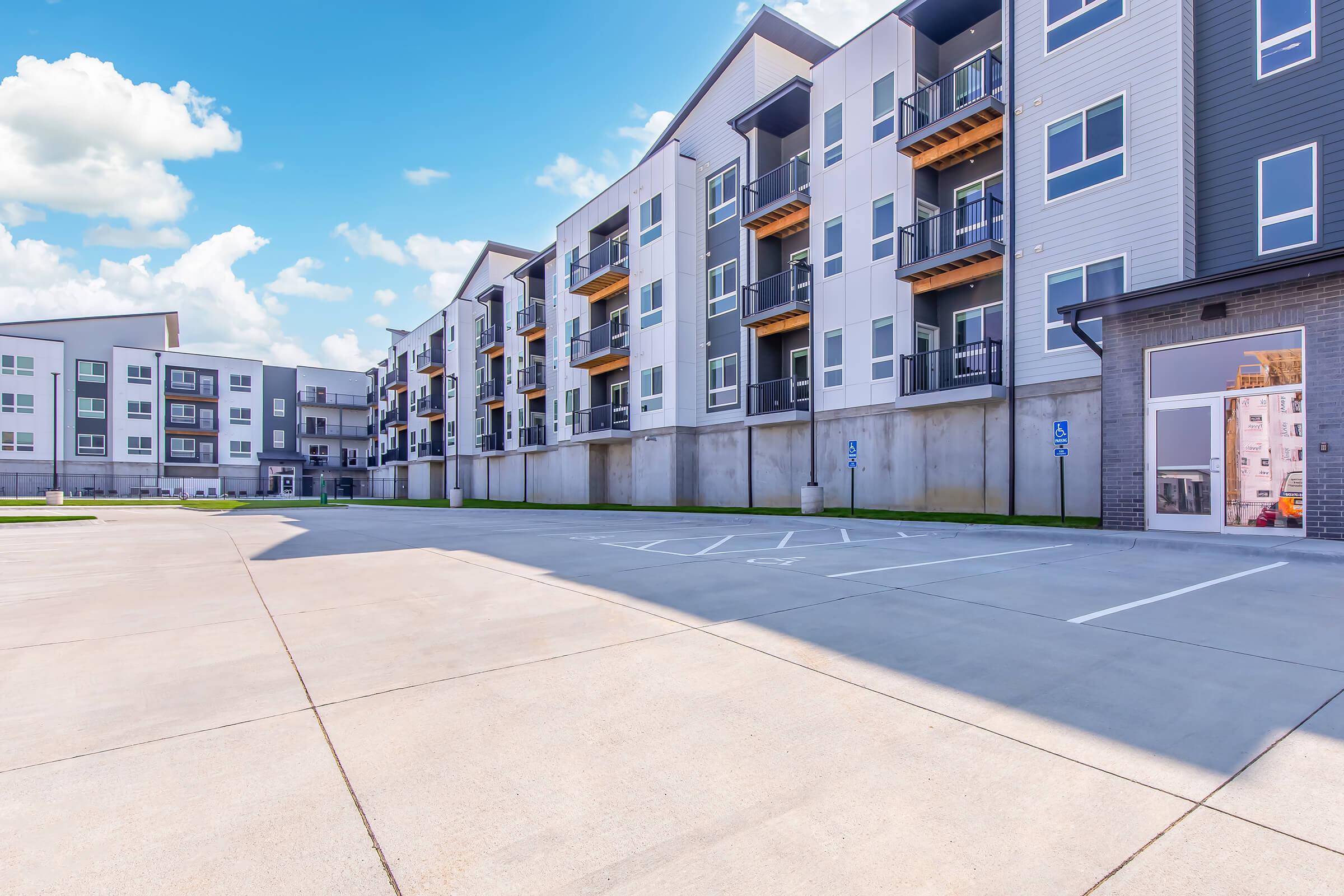 View of a modern apartment complex featuring several multi-story buildings with balconies. The foreground shows a paved parking area with marked spaces, while the backdrop consists of a blue sky with scattered clouds. The buildings have a contemporary design with a mix of colors and materials.