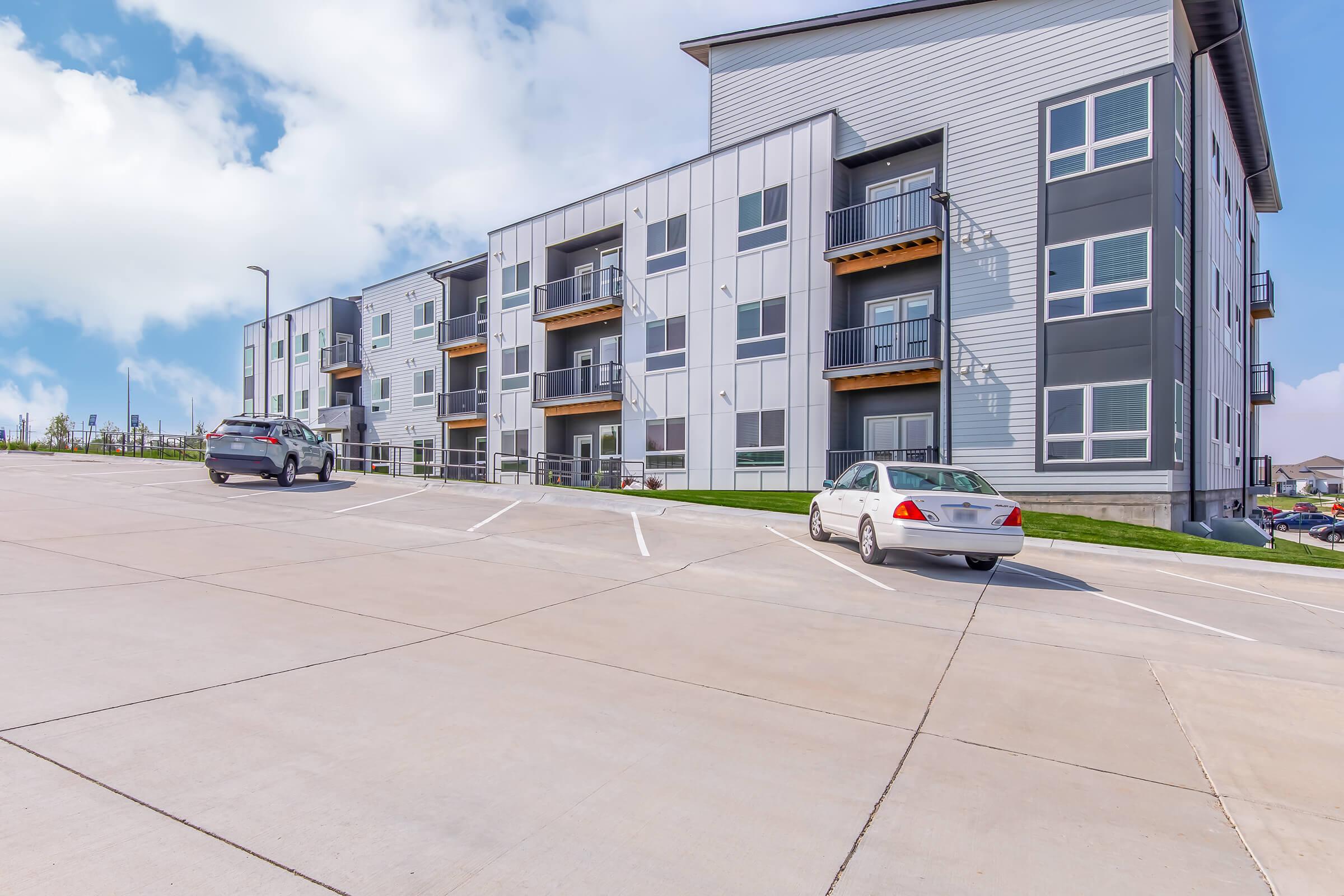 Modern apartment building with multiple units featuring balconies, set against a blue sky with fluffy clouds. The foreground contains a paved parking lot with two cars parked along the side. The architecture is contemporary, with a blend of gray and white panels.