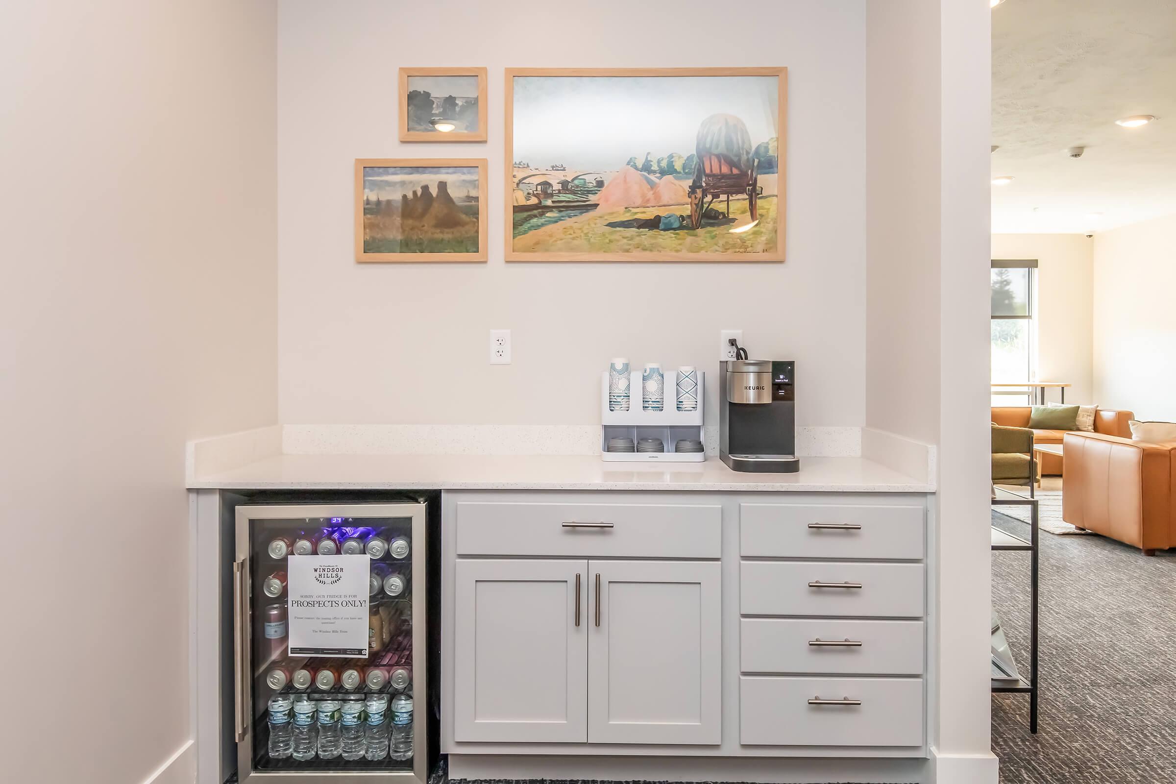 A modern kitchenette featuring a countertop with a coffee machine and several water bottles. Below, a fridge displays more bottled water. The wall is adorned with framed artwork depicting landscapes and historical scenes. The overall design is clean and contemporary, with neutral colors.