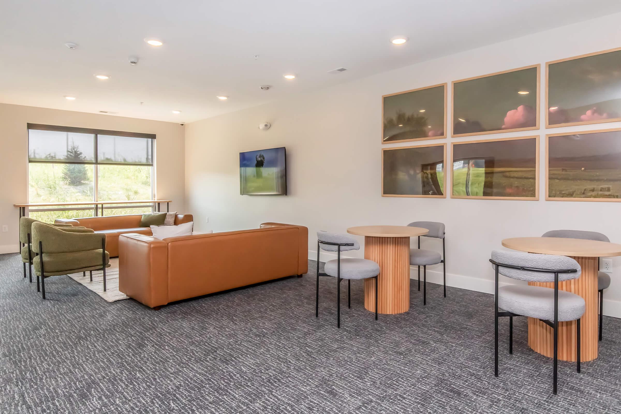 A modern lounge area featuring two sofas, a TV on the wall, and two round tables surrounded by chairs. Large windows allow natural light, and there are framed landscape artworks on the walls. The decor is contemporary, with a neutral color palette and a cozy atmosphere.