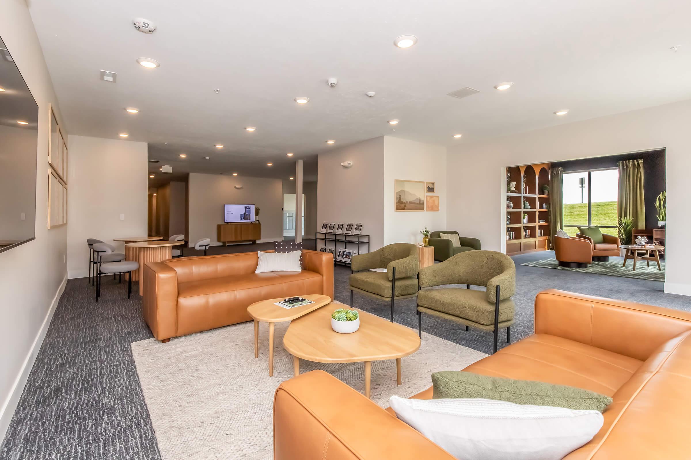 A modern lounge area featuring brown leather sofas, green armchairs, and wooden coffee tables. Soft lighting illuminates the space, with a large TV on one wall and a cozy reading nook in the background. A neutral-toned rug adds texture, creating a warm and inviting atmosphere.