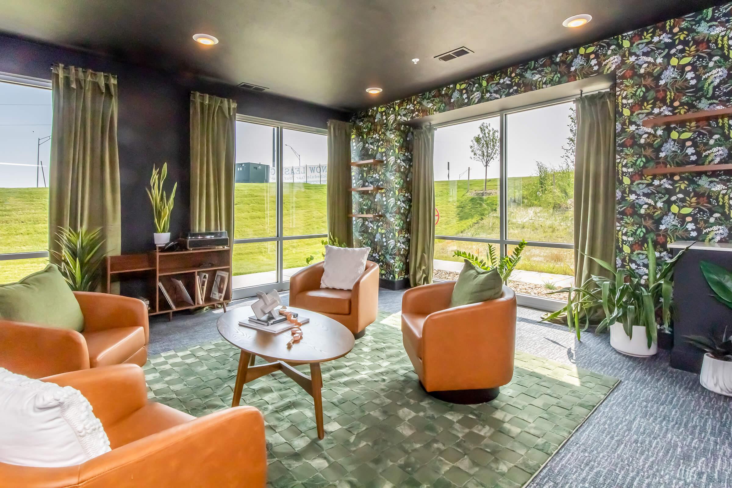 A cozy living area featuring two leather armchairs, a green area rug, and a round coffee table. The walls are adorned with floral wallpaper, and large windows provide natural light and a view of greenery outside. Decorative plants and a wooden shelf add warmth to the space.