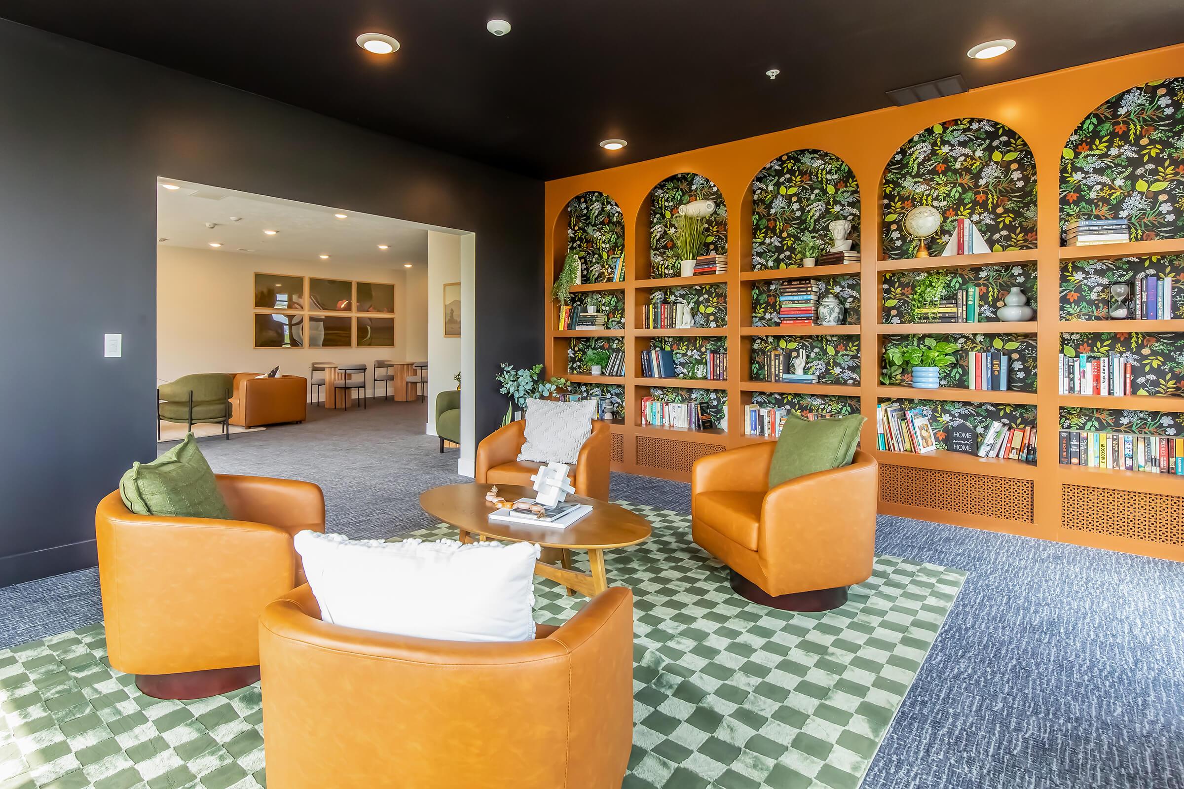 Cozy modern reading area featuring orange-accented walls, a decorated bookshelf filled with books, and four comfortable brown chairs surrounding a round table. The floor is covered with a green patterned rug, and there are decorative plants adding a touch of nature to the space.