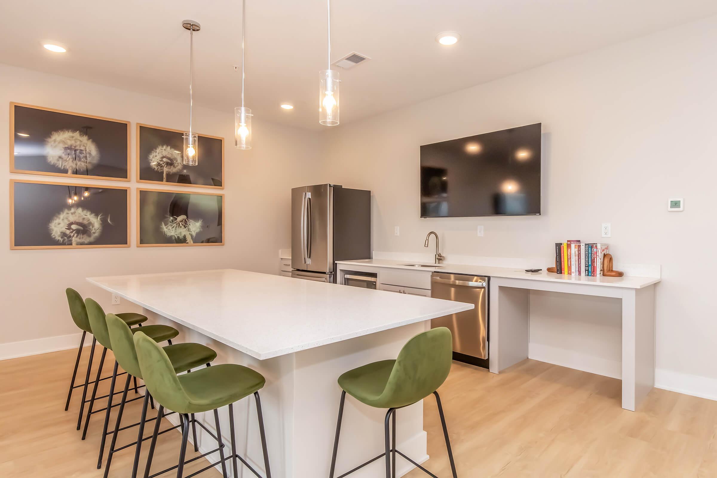 Modern kitchen featuring a large island with green bar stools, stainless steel appliances, and a flat-screen TV mounted on the wall. There are four framed floral images on the wall, and the space is well-lit with pendant lights. The flooring is light wood, creating a bright and inviting atmosphere.