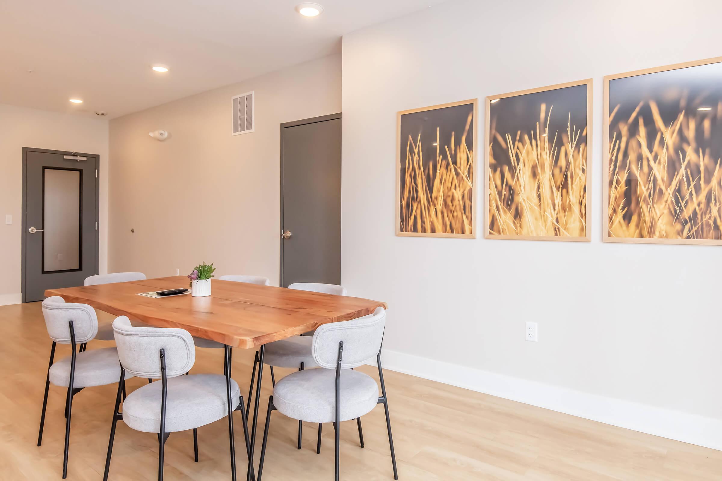 A modern dining area featuring a wooden table surrounded by six gray upholstered chairs. On the wall, there are three framed art pieces depicting abstract images of tall grass. The room has a light, neutral color scheme with warm lighting, creating a welcoming atmosphere.