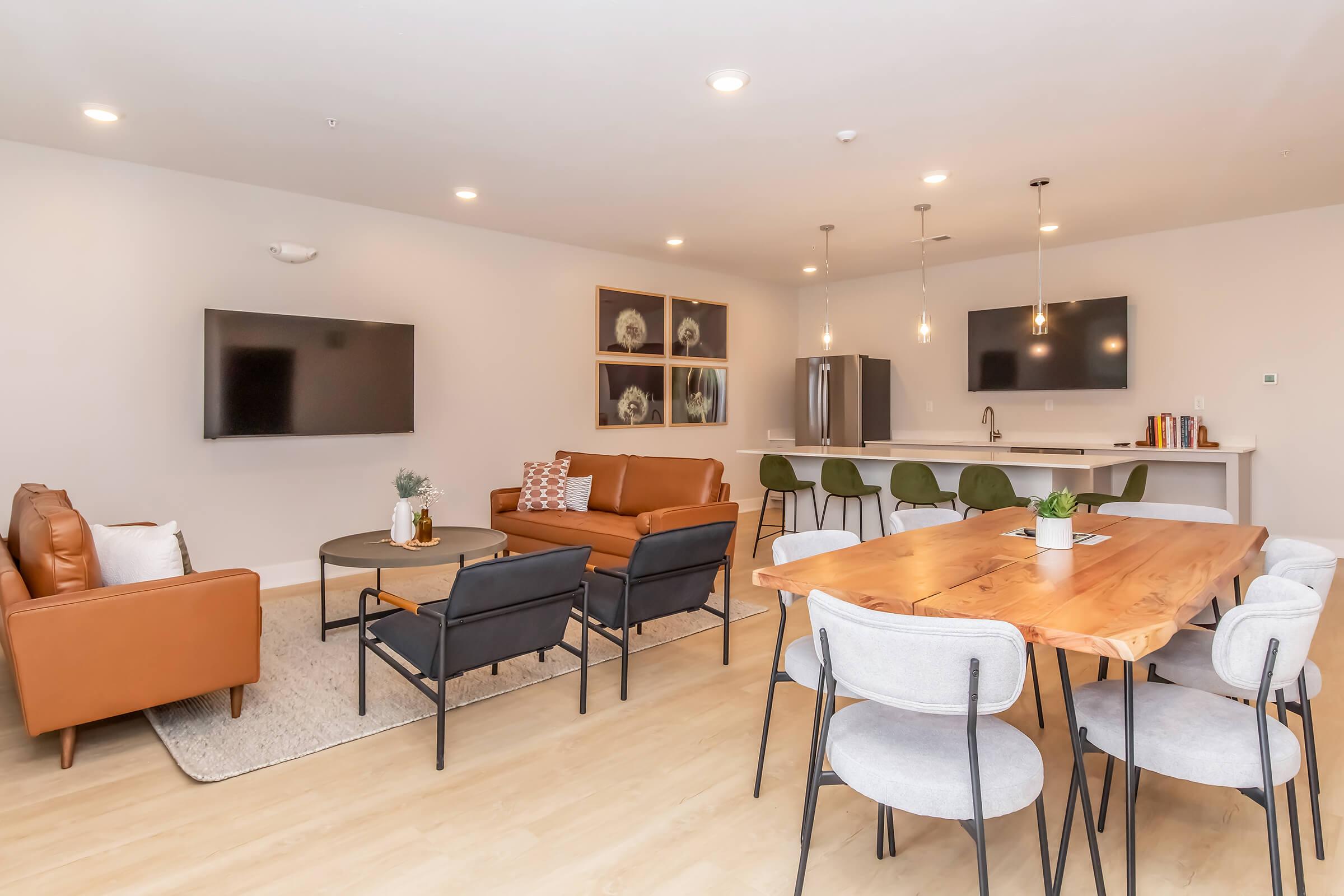 Modern communal living area featuring a cozy seating arrangement with leather and fabric chairs, a wooden dining table, and a stylish kitchen. The space includes large wall-mounted TVs, a neutral color palette, and decorative elements like plants and artwork, creating a welcoming atmosphere.