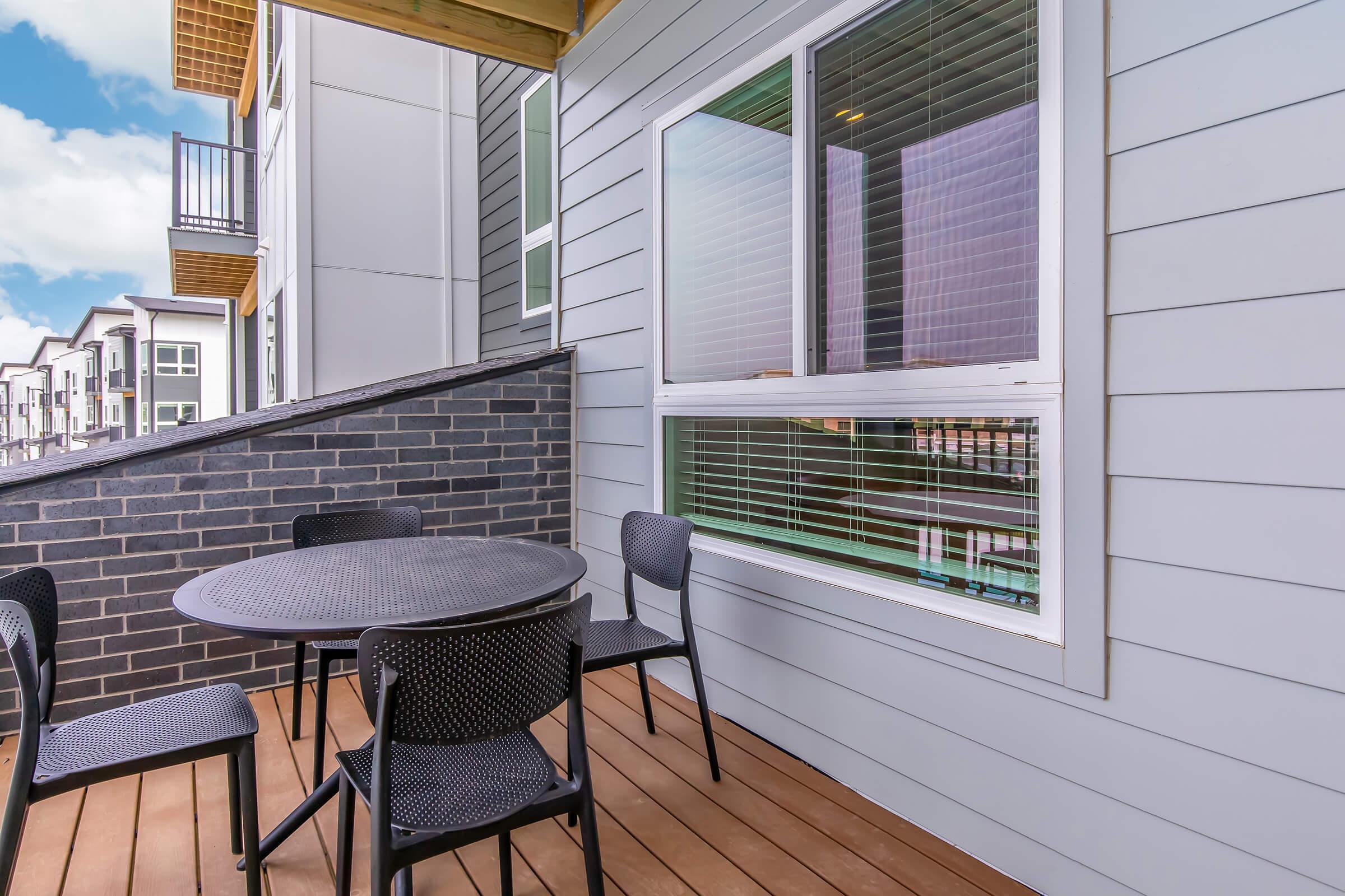 A balcony with a circular black table and four chairs, situated on a wooden deck. The balcony features a gray wall and a window with blinds. In the background, additional apartment buildings can be seen, suggesting a modern living space. The sky is partly cloudy.