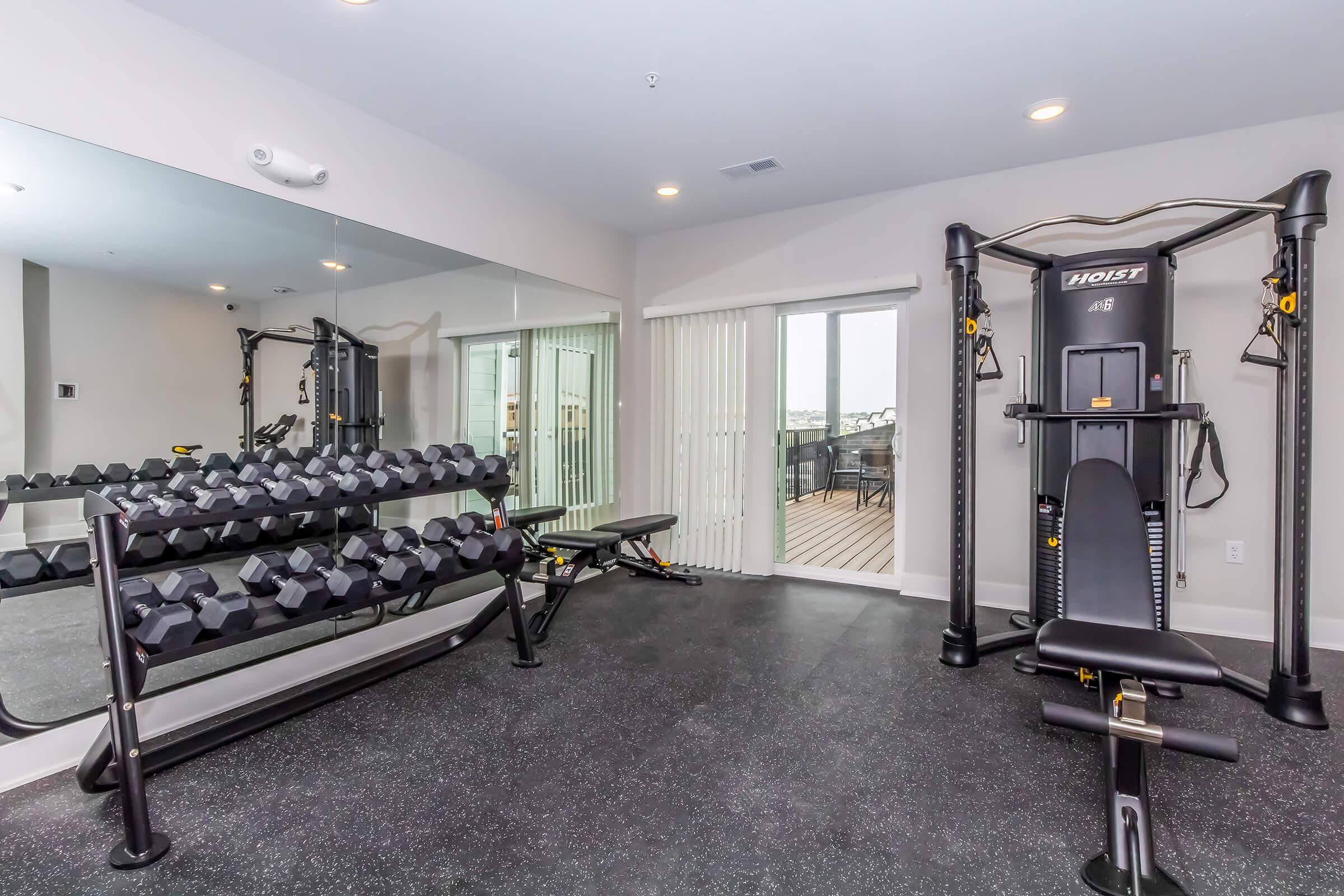 A modern home gym featuring a weight bench, a set of dumbbells on a rack, and a multi-use weight machine. Large mirrors and sliding glass doors provide ample lighting and an outdoor view. The floor is covered with black rubber flooring for safety and comfort.