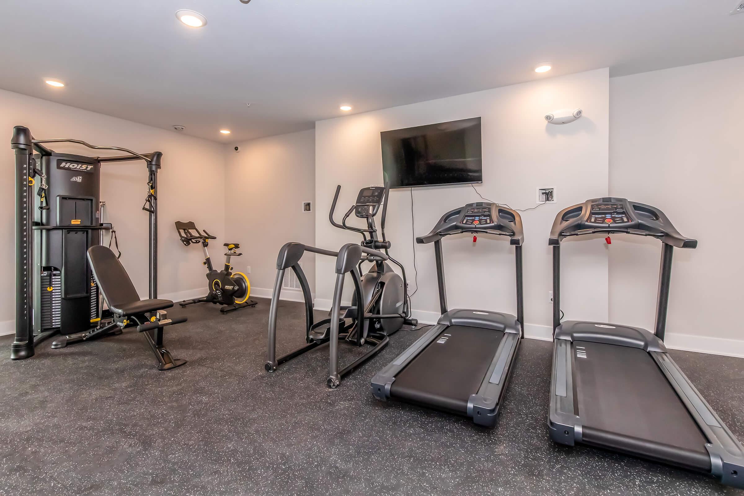 A modern gym featuring two treadmills, an elliptical machine, a stationary bike, and a strength training machine. There is a flat-screen TV mounted on the wall, and the floor is covered in black rubber flooring, creating a functional workout space.