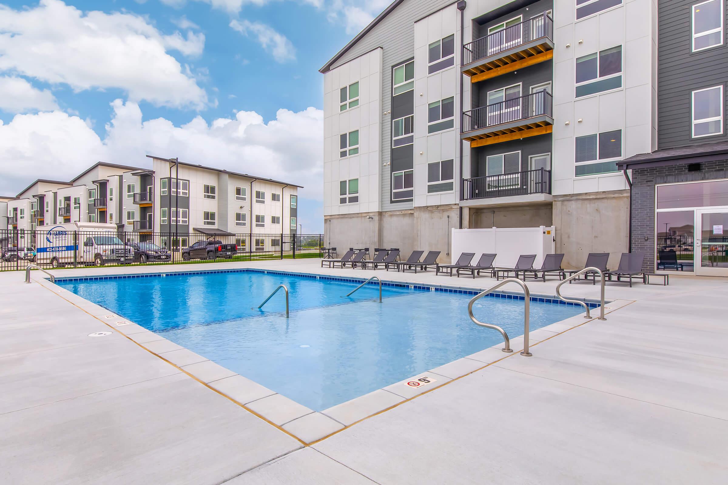 A clean and inviting outdoor swimming pool surrounded by lounge chairs, located at an apartment complex. The pool features steps for easy access, with modern buildings in the background under a bright blue sky.
