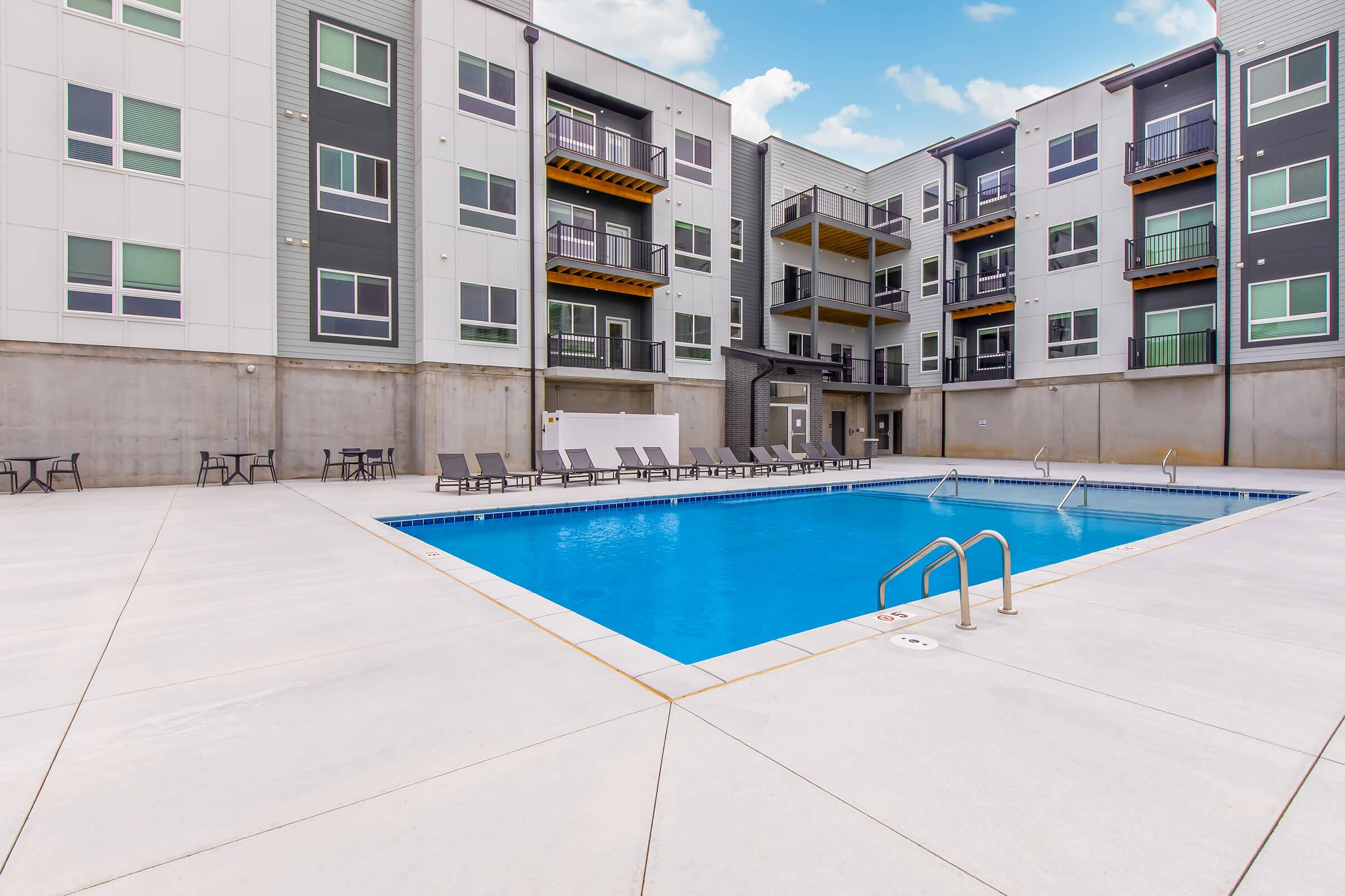A modern outdoor swimming pool surrounded by a concrete patio. Lounge chairs are arranged around the pool area, with residential buildings featuring large windows in the background. The sky is partly cloudy, and the setting has a clean, contemporary aesthetic.