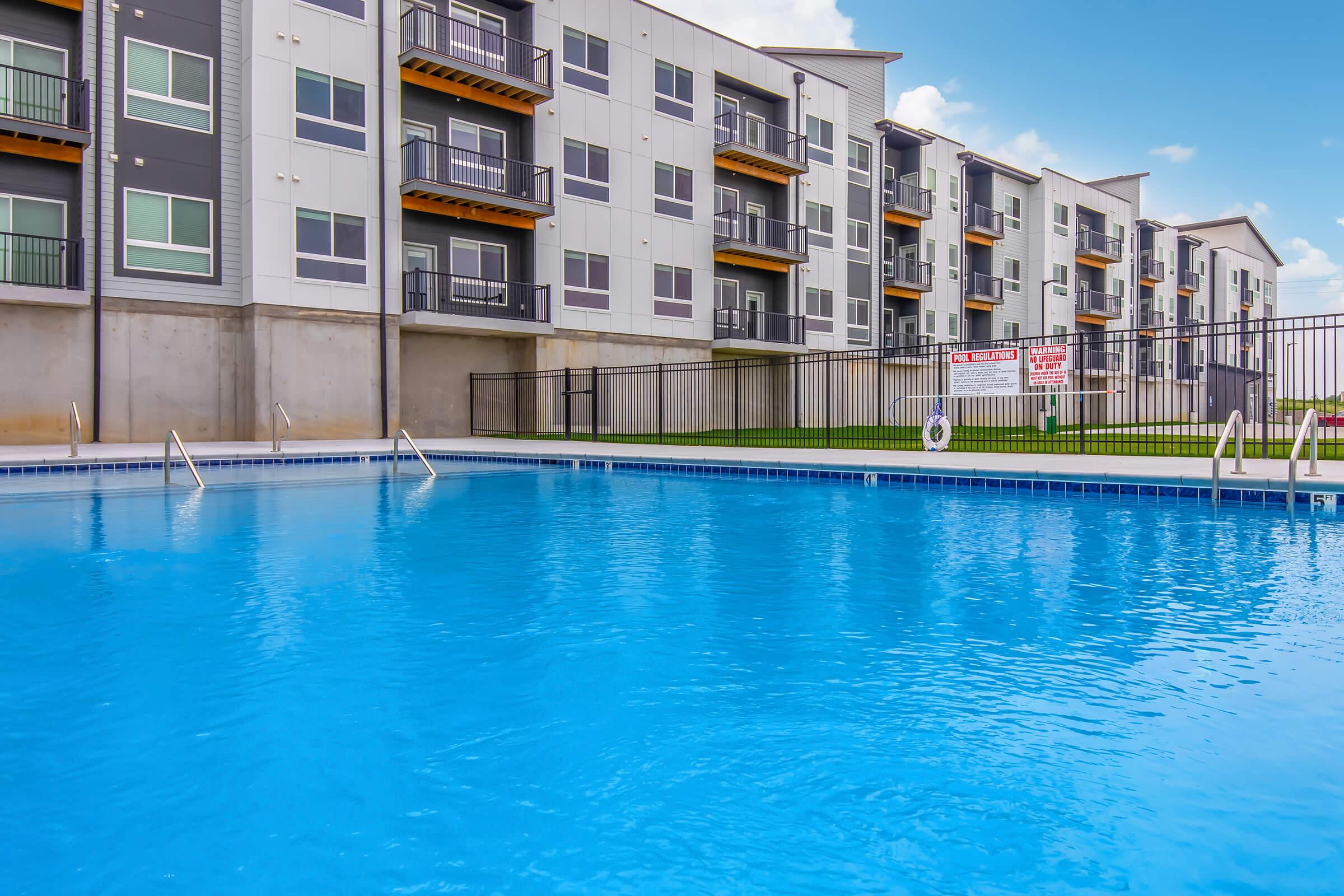 A clear blue swimming pool in the foreground, with steps leading into the water. In the background, multi-story residential buildings featuring balconies and large windows. The area is well-maintained with green grass and a safety sign near the pool. Bright blue sky adds to the inviting atmosphere.