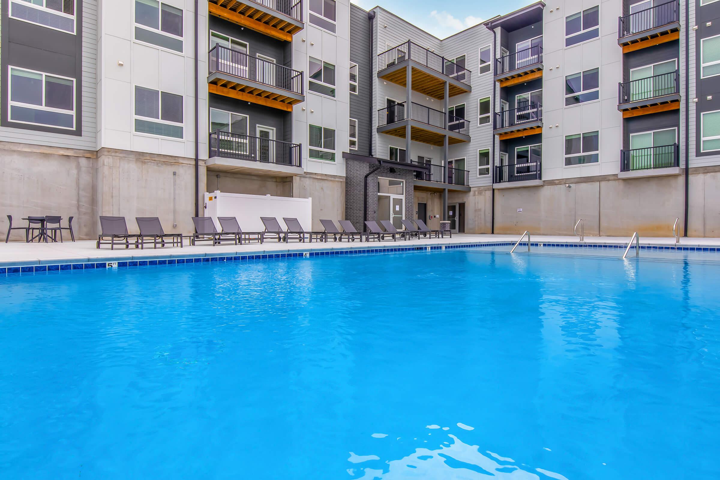A refreshing outdoor swimming pool surrounded by lounge chairs, with modern apartment buildings in the background. The water is clear and blue, and the atmosphere is inviting for relaxation and leisure.