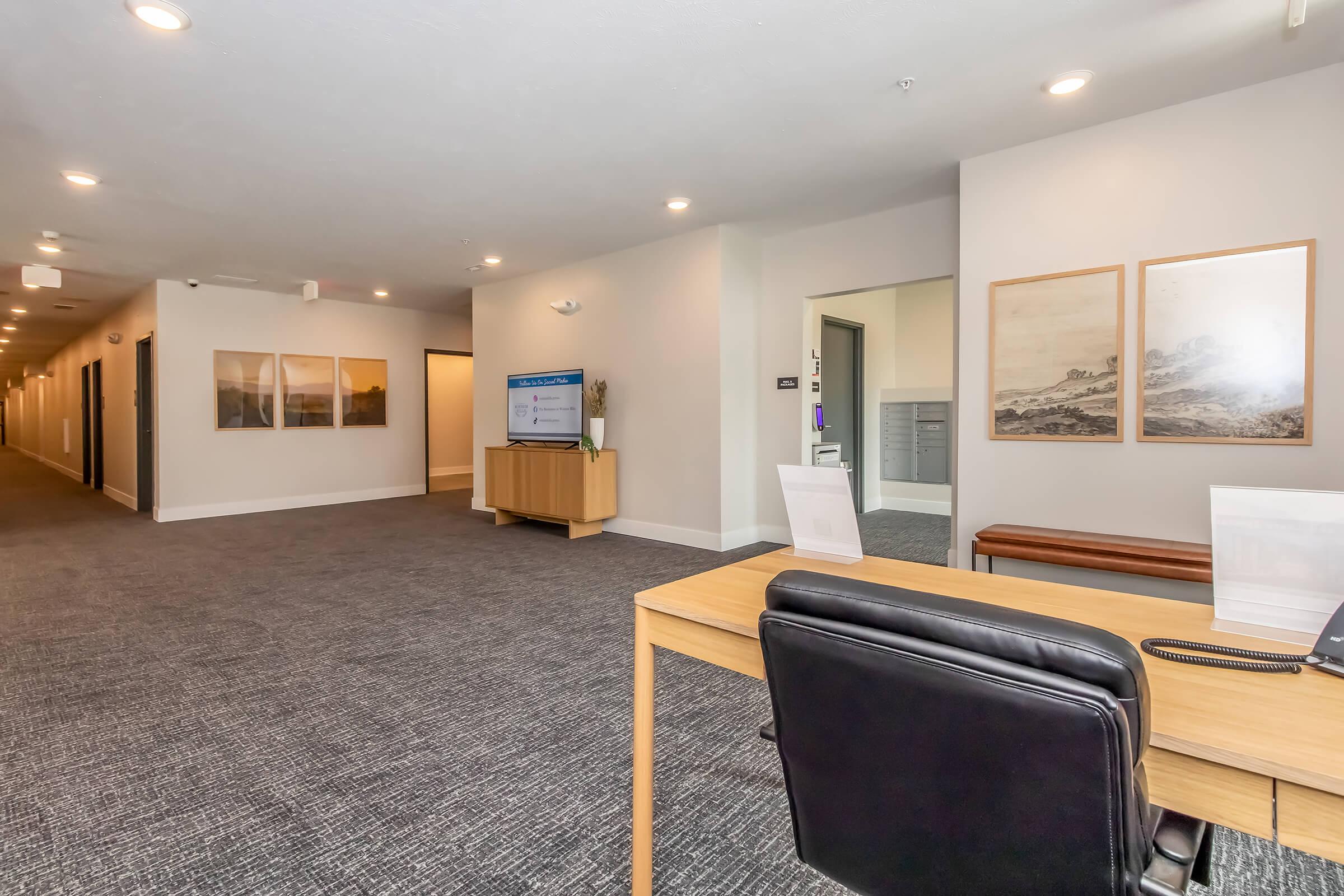 Interior view of a modern hallway with gray carpet and neutral walls. On the right, a reception desk with a black chair and a phone. Two framed pictures hang on the wall behind the desk. A large screen is visible on the left, displaying information. The hallway extends further into the background with additional doors.
