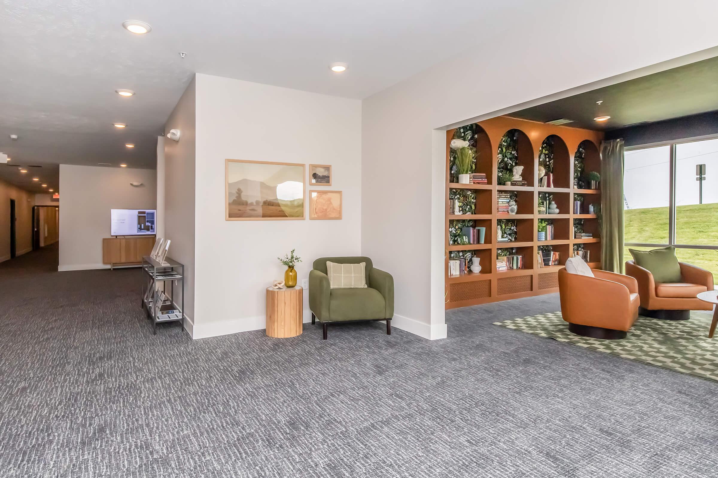 Interior view of a modern hallway featuring a cozy seating area with an armchair and a small table, alongside a bookshelf against the wall, decorated with plants. The space has neutral tones and a carpeted floor, with soft lighting creating a welcoming atmosphere.