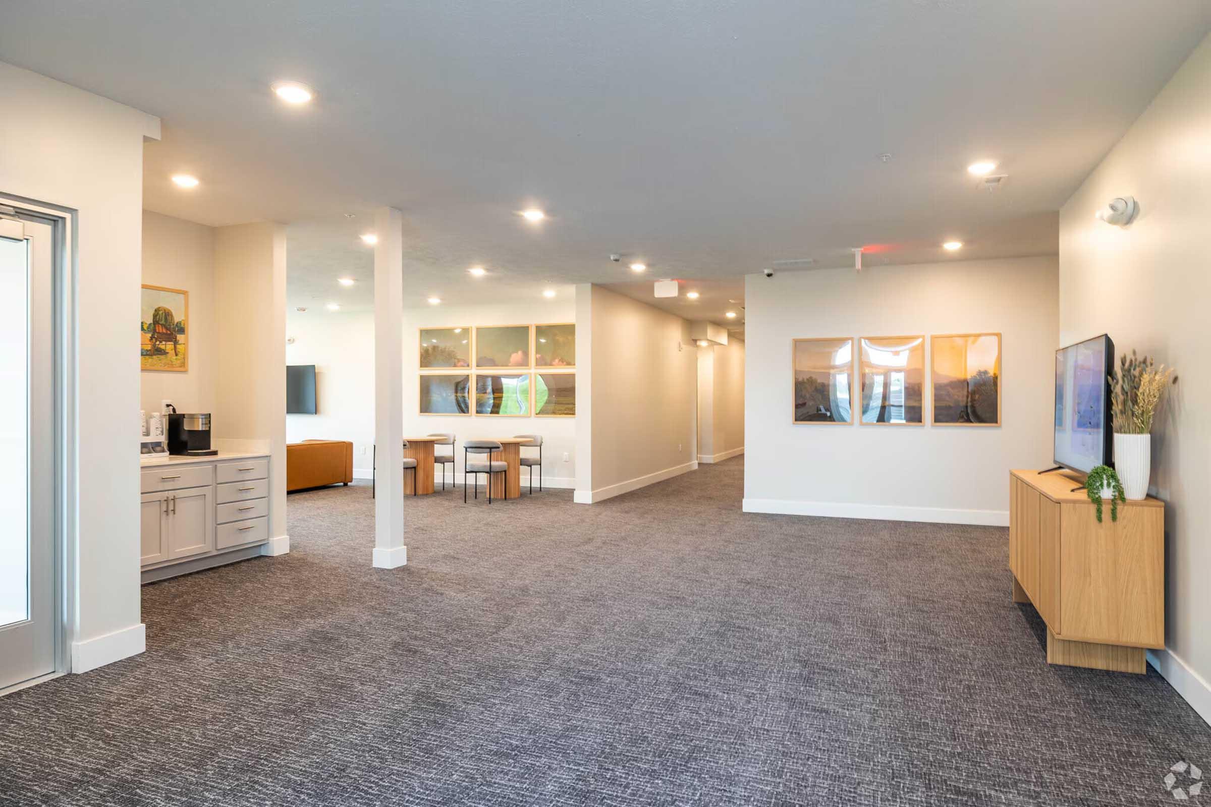 A spacious indoor area with gray carpet flooring and neutral-colored walls. To the left, there is a kitchenette with a coffee machine and seating area. In the center, a dining table with chairs. On the right, a TV mounted on the wall above a wooden console, with artwork hung on the adjacent wall. Soft lighting creates a welcoming atmosphere.