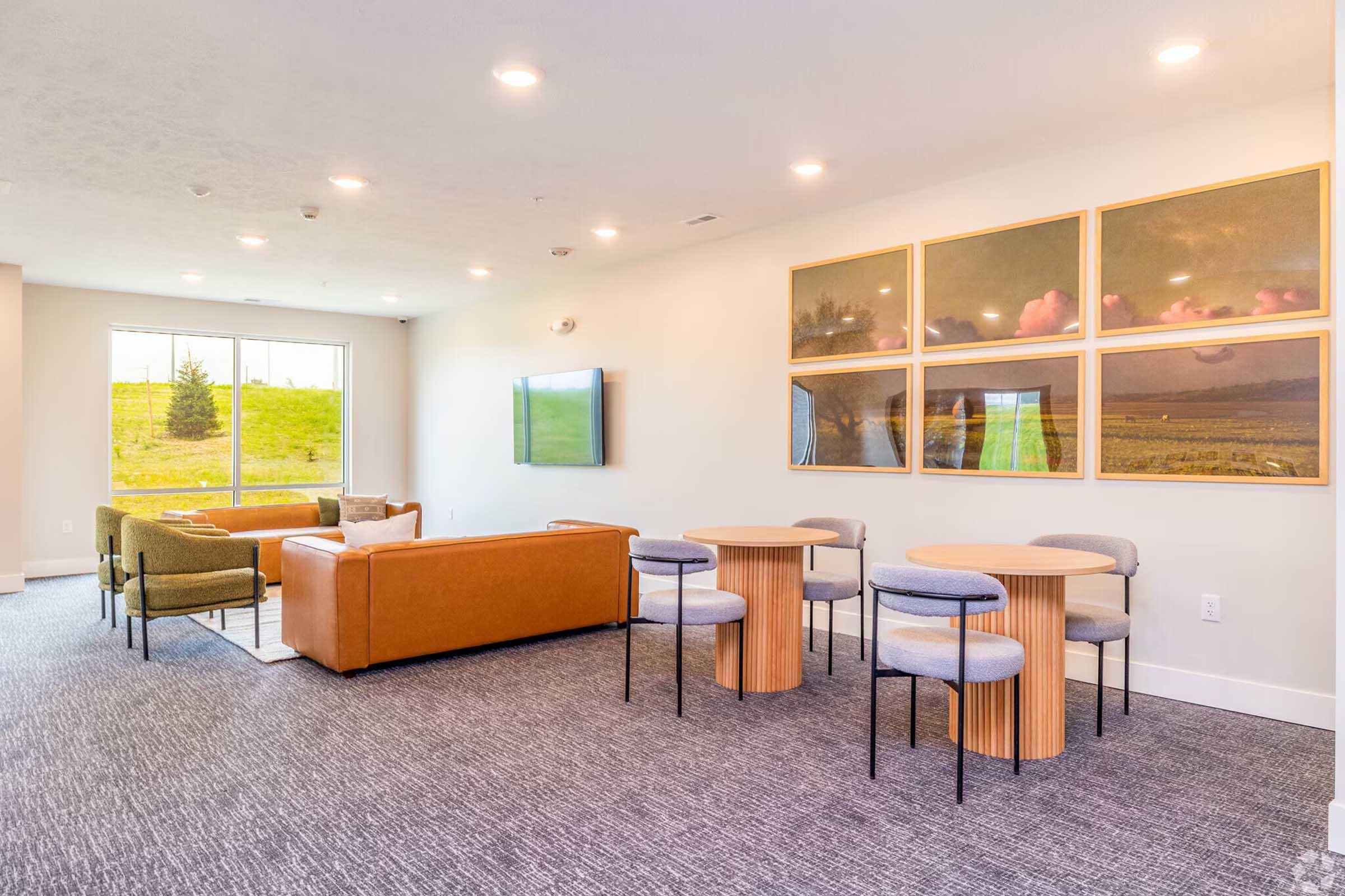 A well-lit lounge area featuring modern furniture, including a brown leather sofa, two green chairs, and round tables with gray chairs. A large window overlooks a green landscape, and there are framed pictures on the wall. The space has a cozy and inviting atmosphere, suitable for relaxation or socializing.