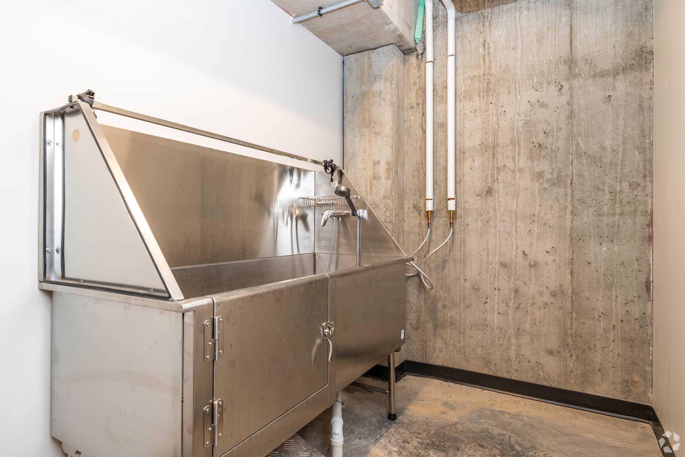 A stainless steel utility sink with a hinged lid, situated against a concrete wall. The room features exposed pipes and a simple, industrial design. The floor is bare and slightly textured, enhancing the utilitarian feel of the space.