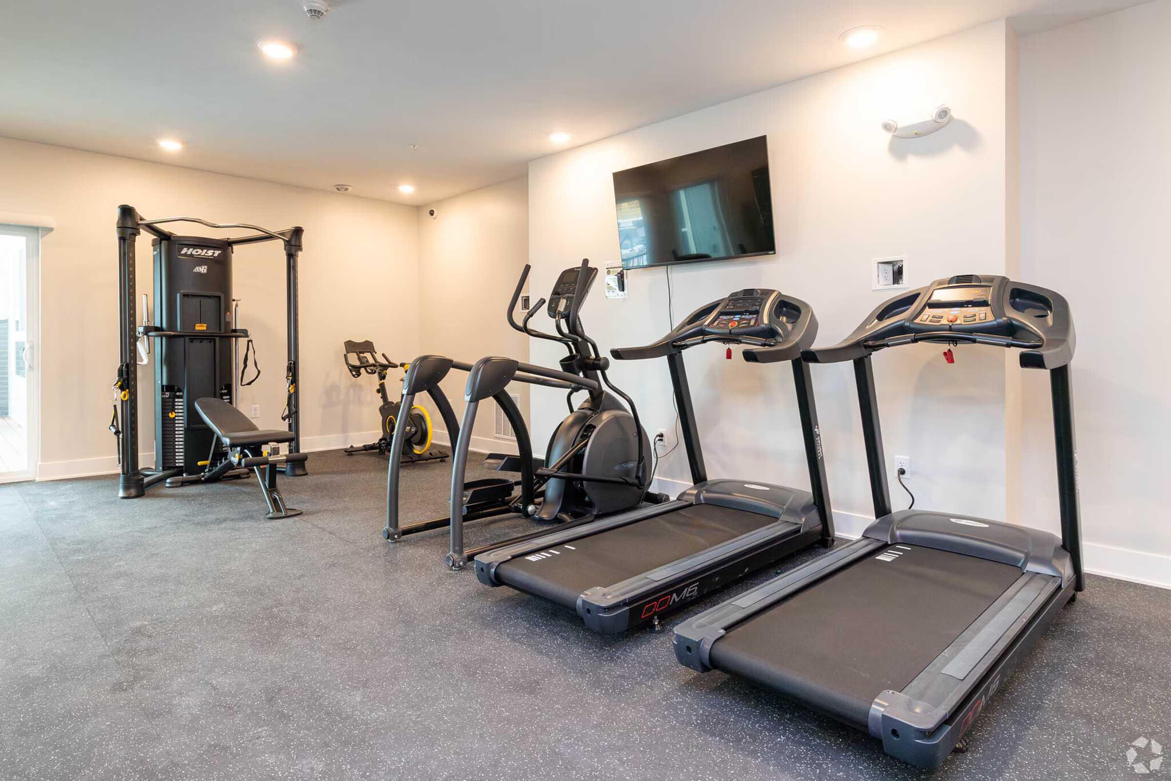 A modern gym interior featuring several exercise machines, including two treadmills, an elliptical, and a multi-station weight machine. The room has a wall-mounted TV, ample natural light, and a rubber floor, creating a functional and inviting workout space.