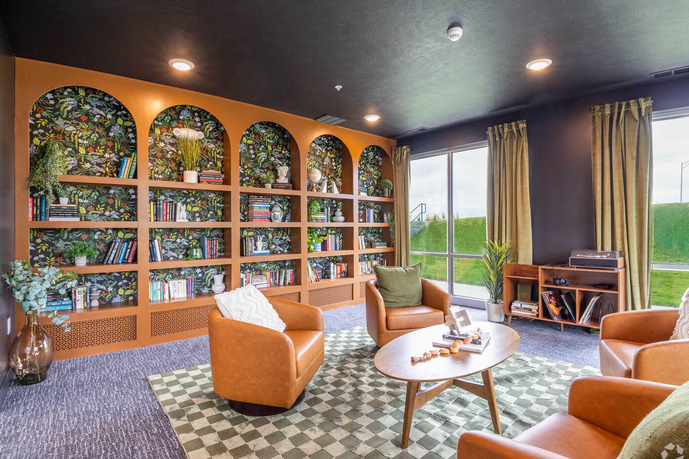 A cozy reading nook featuring two brown leather armchairs and a round wooden coffee table. Behind the seating area is a bookshelf with a green leaf pattern, filled with various books and decorative plants. Large windows let in natural light, and there are green curtains and a patterned rug on the floor.