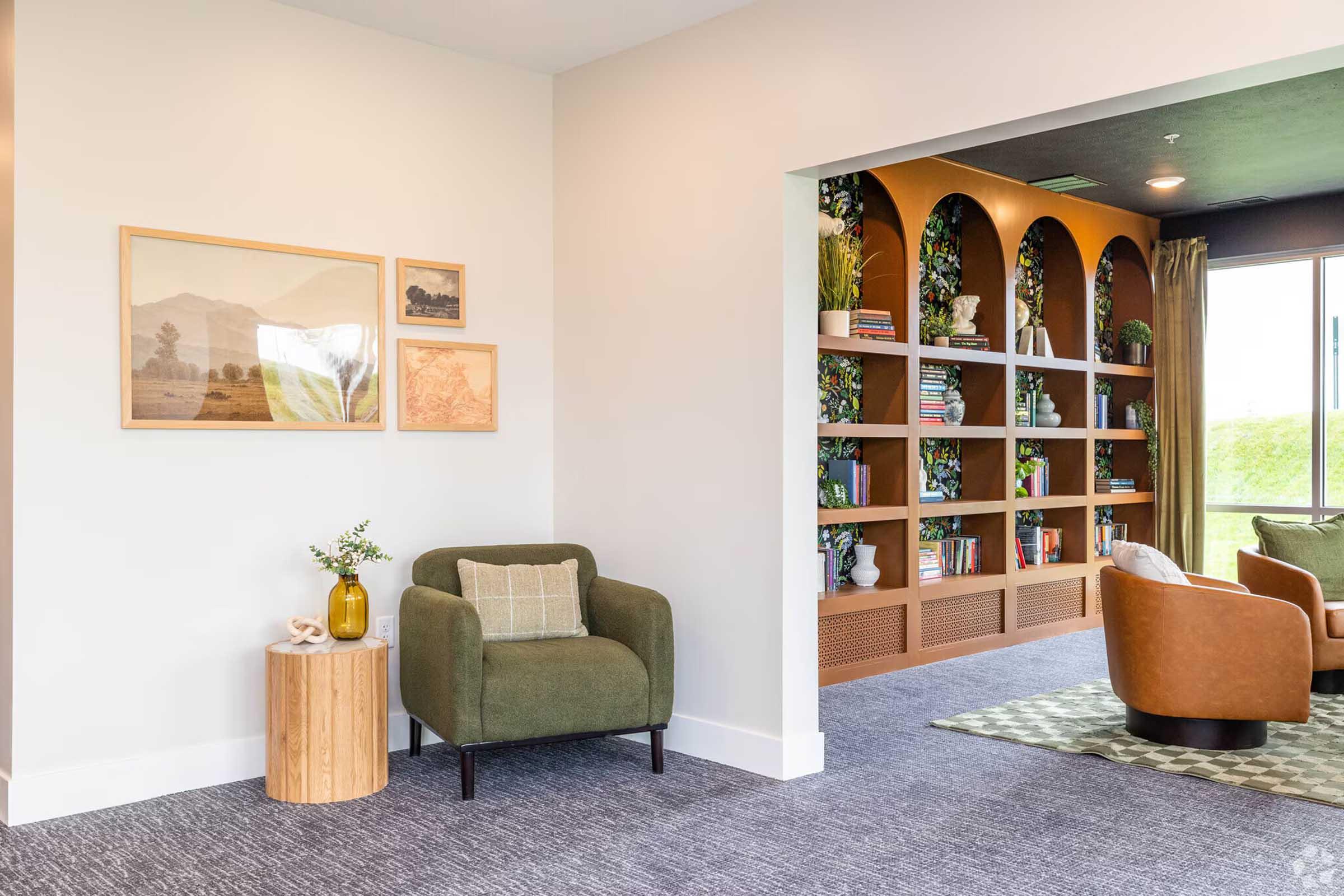 A modern room with a cozy seating area featuring two armchairs and a wooden side table adorned with a decorative vase. One wall displays framed artwork, while another wall showcases built-in shelves filled with books and decorative plants. Natural light streams in through a large window.