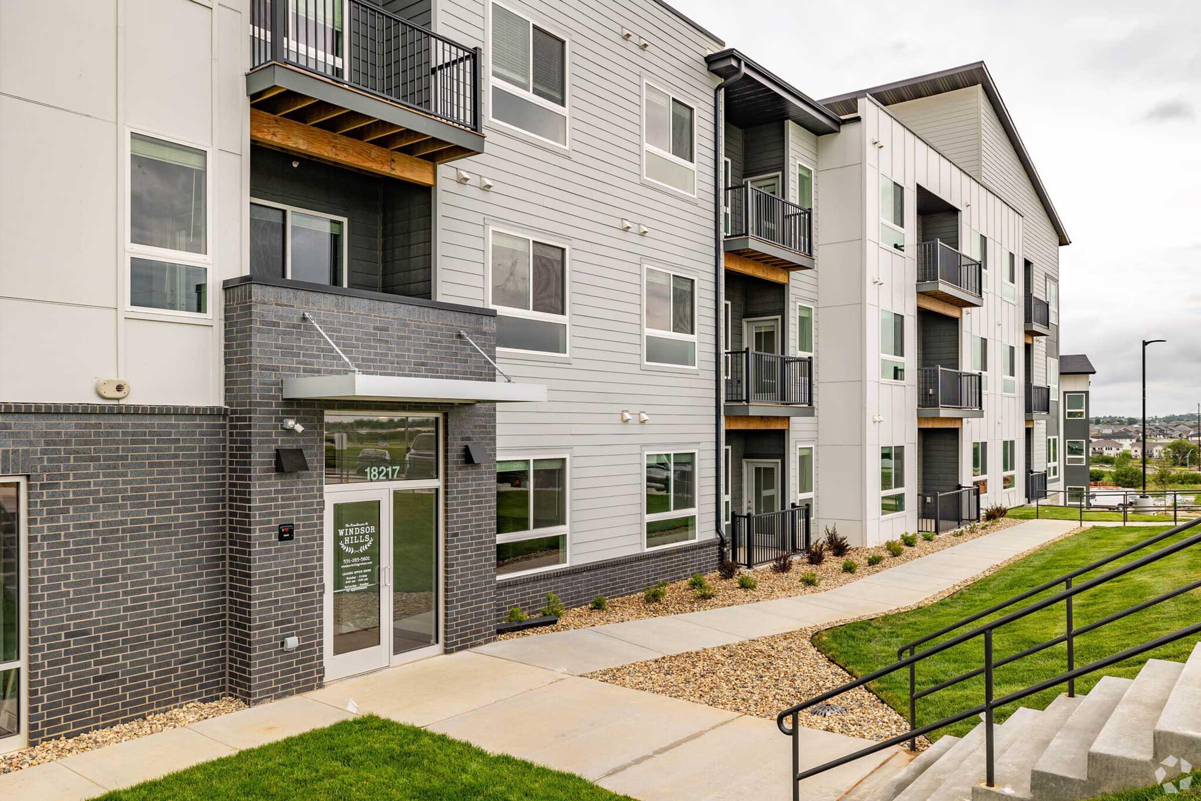 Modern apartment building exterior featuring a mix of gray siding and brick. The entrance is framed by large windows and a canopy. There are balconies on the upper floors, and a landscaped pathway leads to the entrance. The surrounding area includes green grass and decorative stone elements.