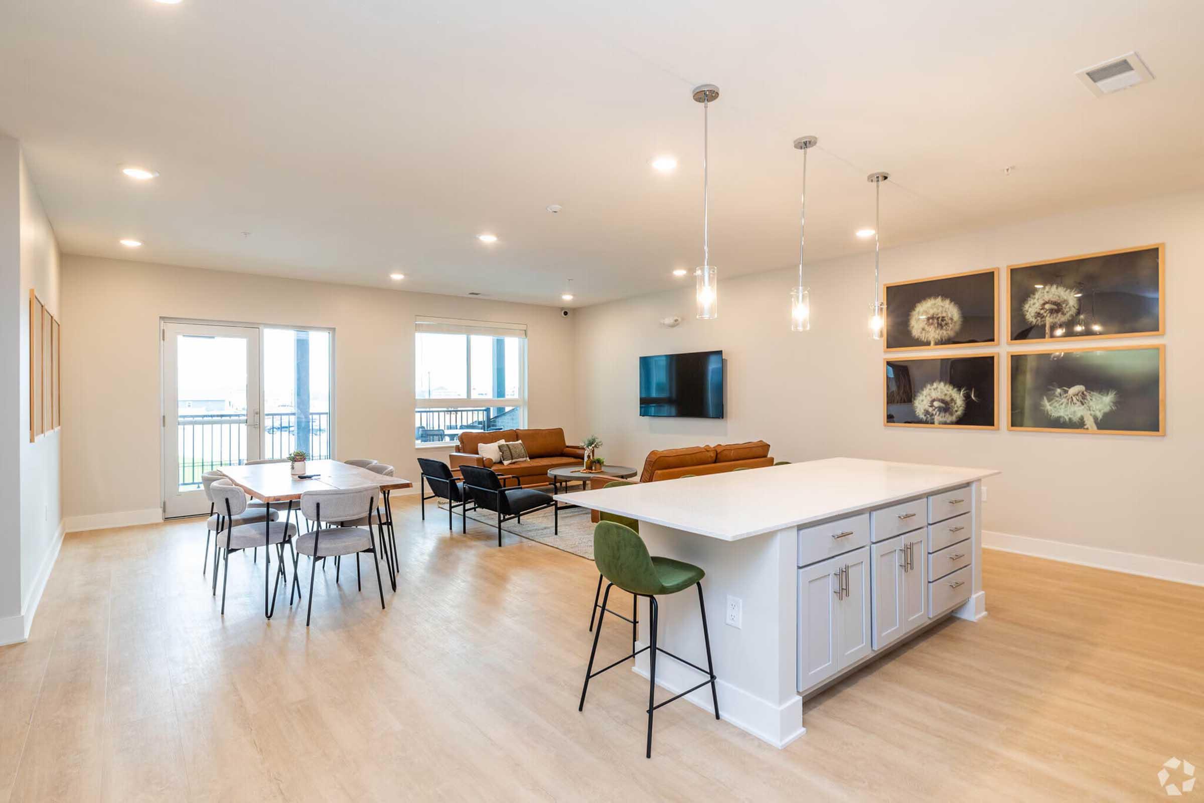 A modern, open-concept living area featuring a light wood floor, a stylish kitchen island with bar stools, a dining table, and a cozy seating area with two sofas. Large windows allow natural light, and decorative wall art adds a contemporary touch.