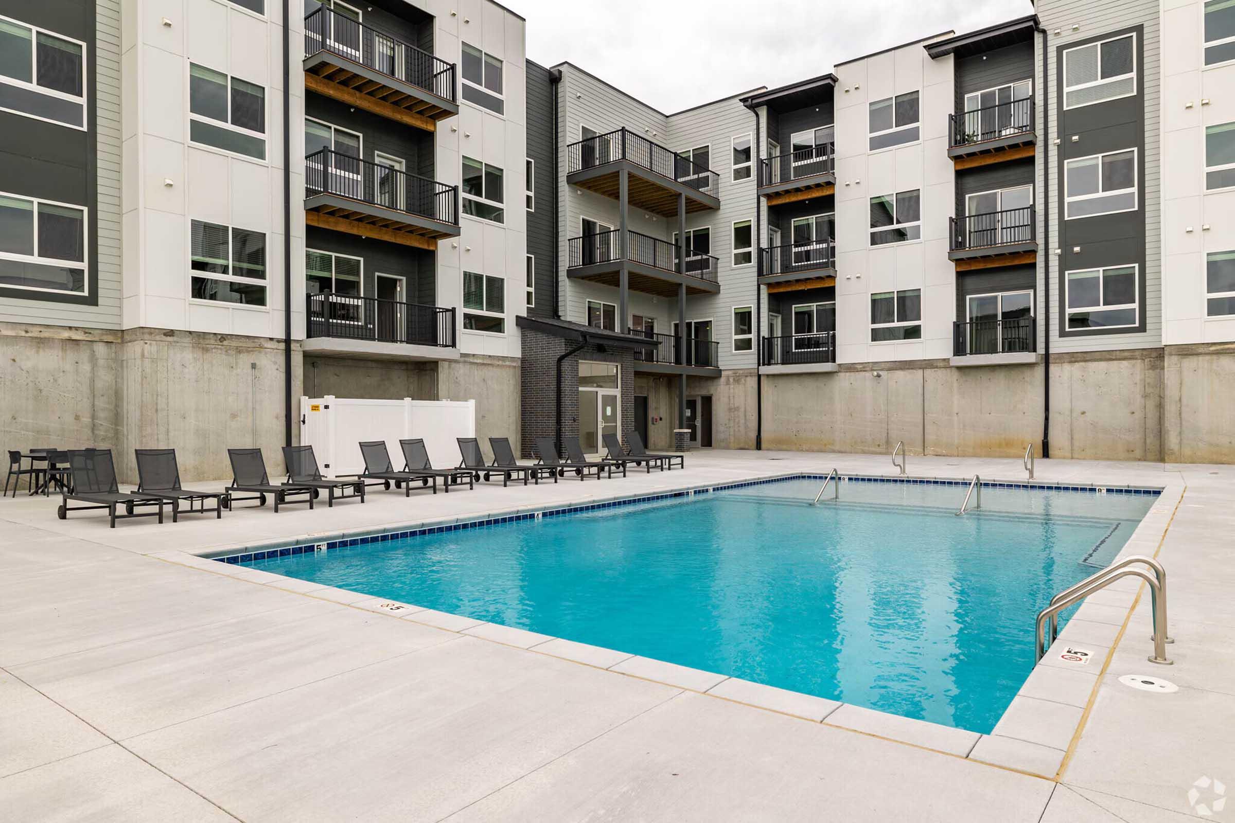 A clean, modern apartment complex featuring a swimming pool in an outdoor area. The pool is surrounded by loungers and has steps for entry, with three-story buildings in the background. The architecture is contemporary, with balconies and large windows. The setting appears inviting and well-maintained.