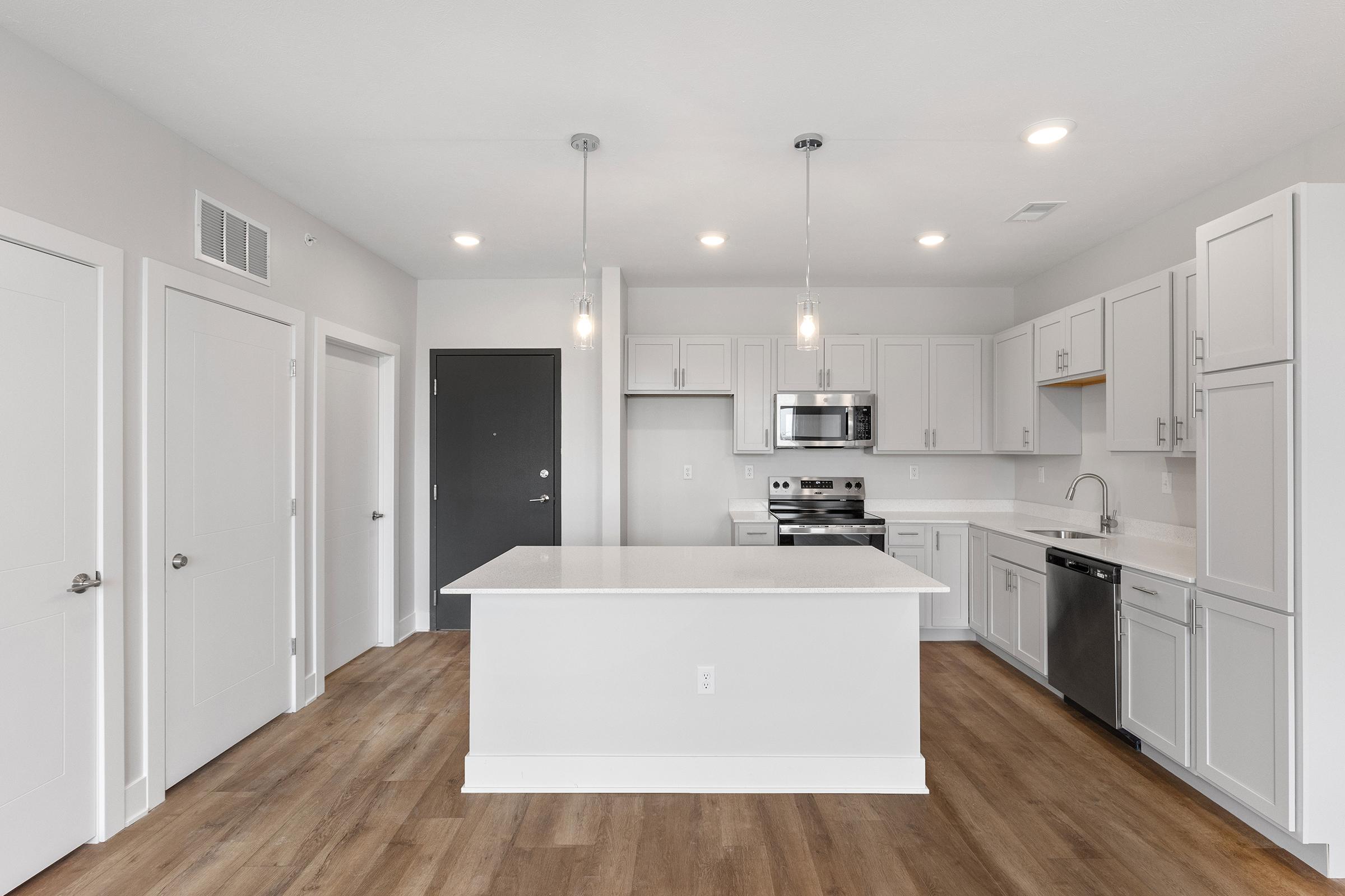 Modern kitchen featuring light-colored cabinetry, stainless steel appliances, and quartz countertops. An island with bar seating is centered in the space, complemented by pendant lighting. The kitchen opens to a bright area with neutral-toned walls and wood-like flooring, enhancing the contemporary feel.