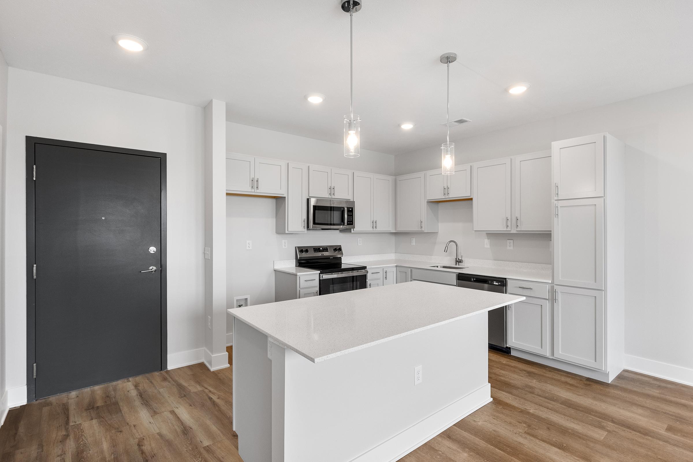 Modern kitchen featuring white cabinetry, stainless steel appliances, a large island with a quartz countertop, and hardwood flooring. Well-lit with pendant lights above the island and an open layout connecting to the living area. The entrance is seen from the left side of the image.