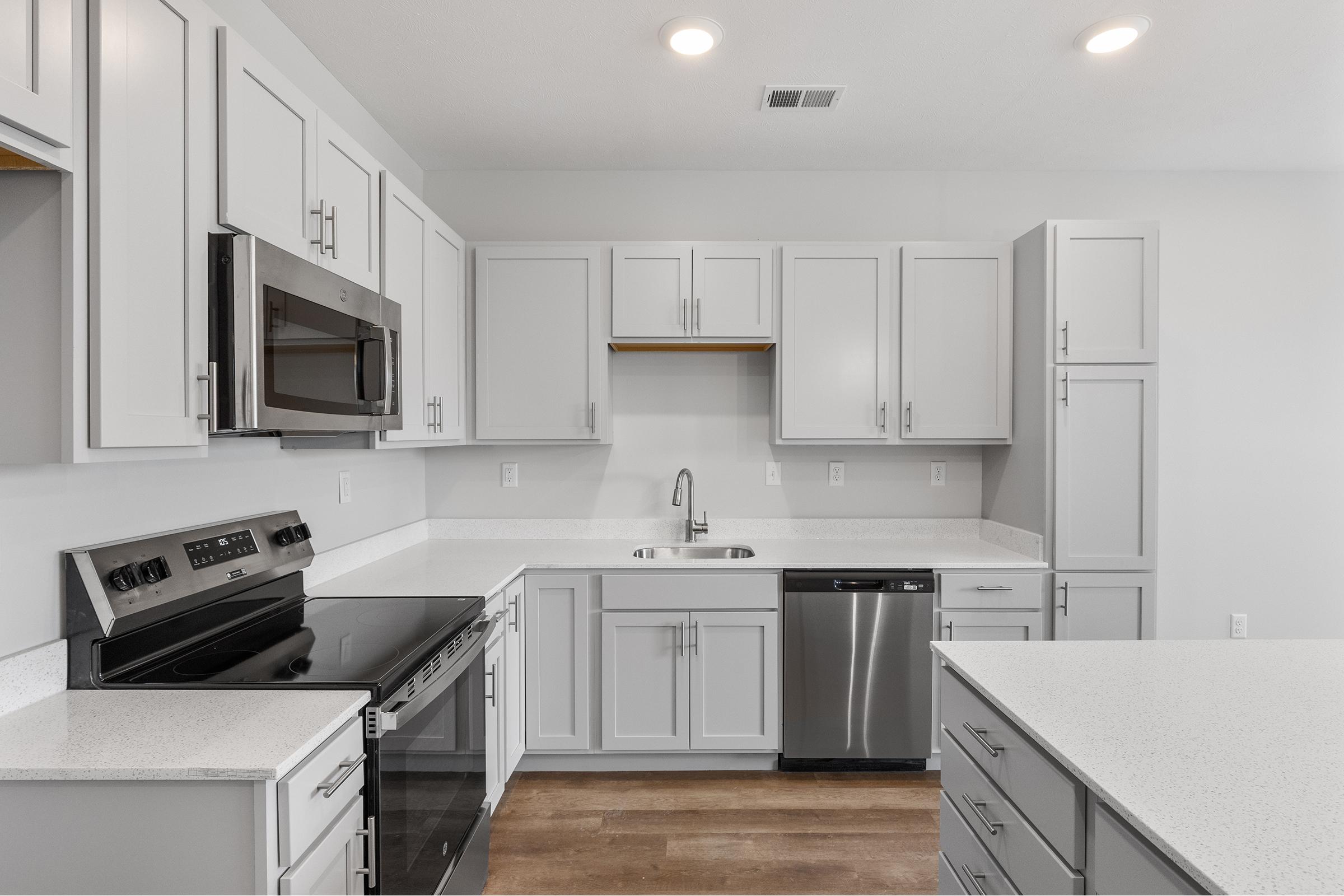 Modern kitchen featuring light gray cabinets, stainless steel appliances including an oven, microwave, and dishwasher. The countertops are white with a subtle pattern, and the floor is a warm wood tone. The space is bright with overhead lighting and a minimalist design.