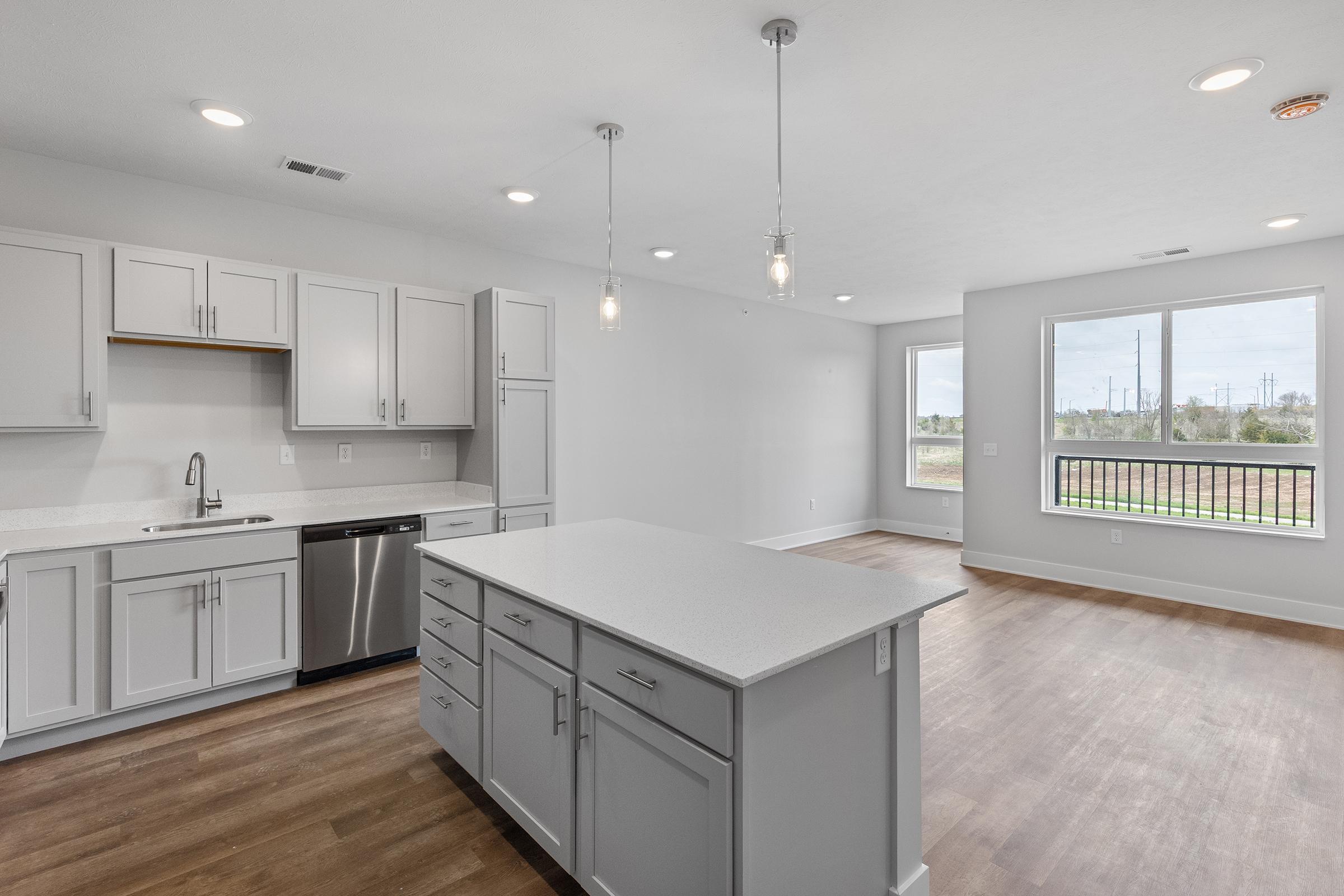 Modern kitchen interior featuring light gray cabinetry, a central island with a white countertop, and stainless steel appliances including a dishwasher. The open layout connects to a bright living area with large windows, allowing for natural light and a view of the outdoor landscape. Wood-like flooring adds warmth to the space.