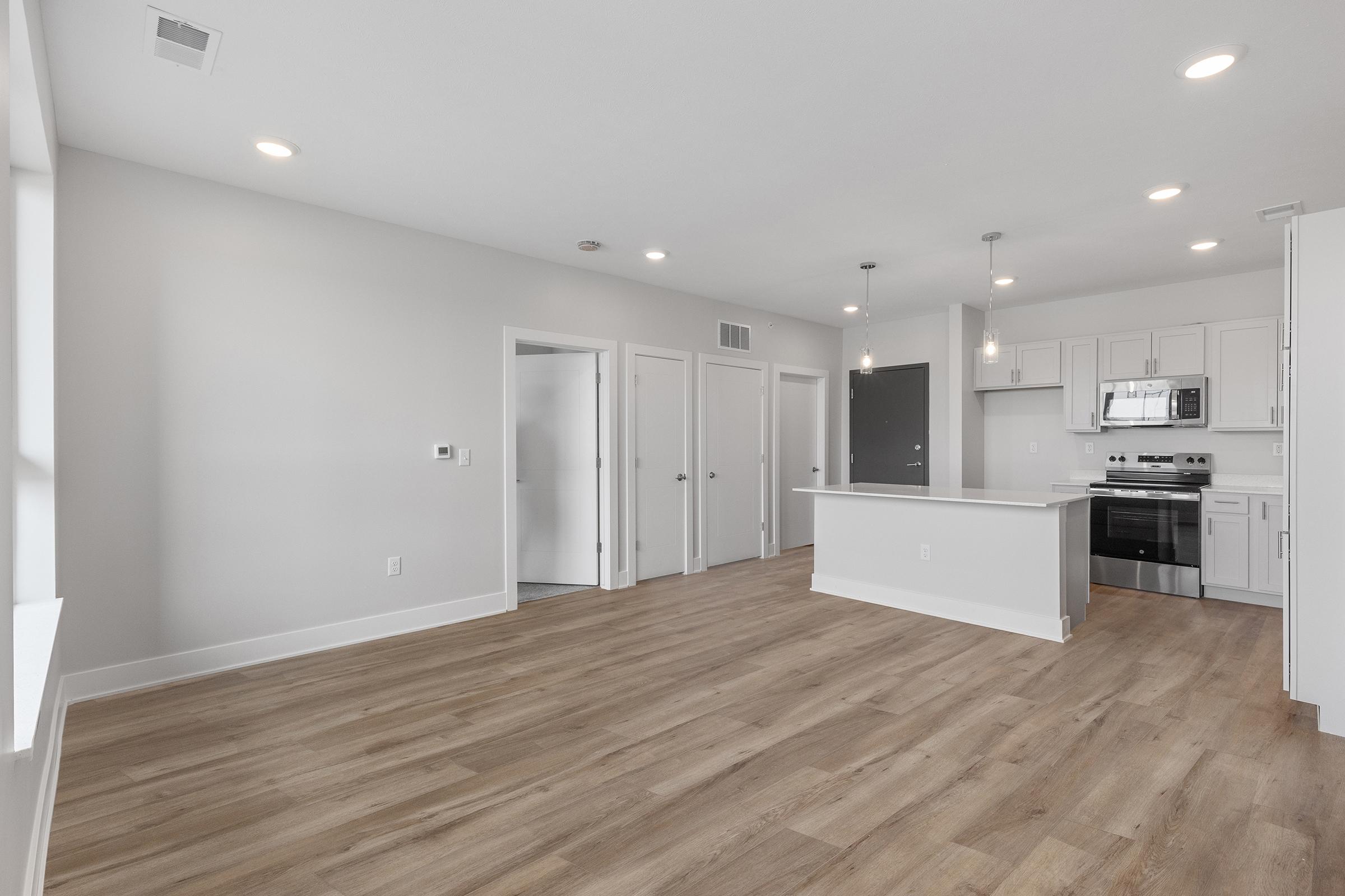 Spacious modern apartment interior featuring light grey walls, hardwood floors, and an open layout. The kitchen includes stainless steel appliances and a central island, while multiple doors lead to bedrooms or utility areas. Large windows provide natural light, enhancing the airy feel of the living space.